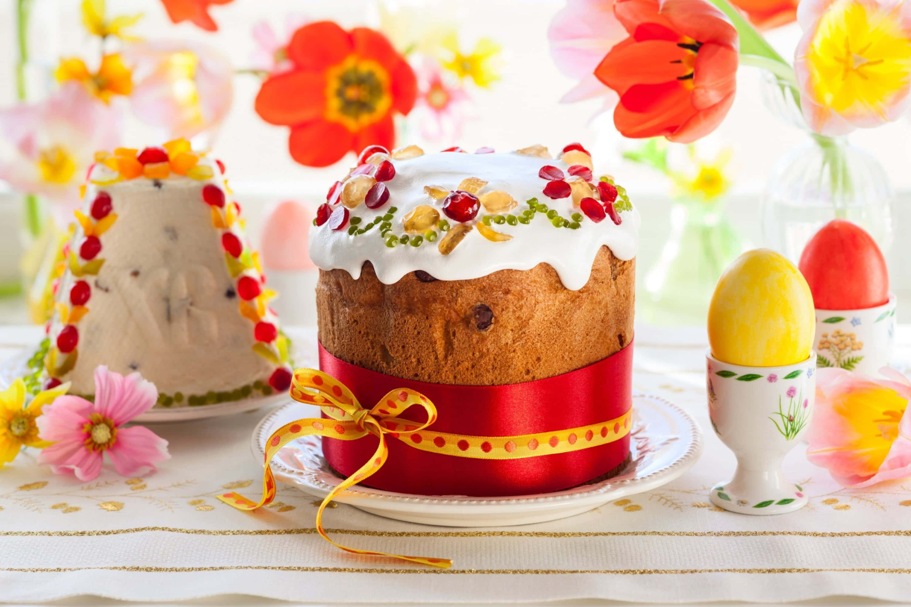 Spring Desserts 桌布 壁紙 — 一個節日復活節蛋糕，飾有紅色漿果和綠色珠子，放在紅色絲帶上，附近有彩色雞蛋和春天花朵。