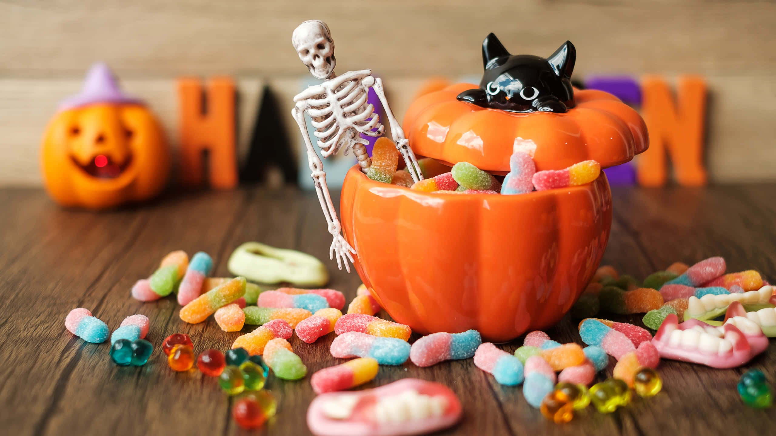Halloween Treats 桌布 壁紙 — 一個節日氣氛的萬聖節場景，木桌上有一個南瓜，裡面裝滿了色彩繽紛的糖果，骷髏和黑貓從南瓜中探出頭來。