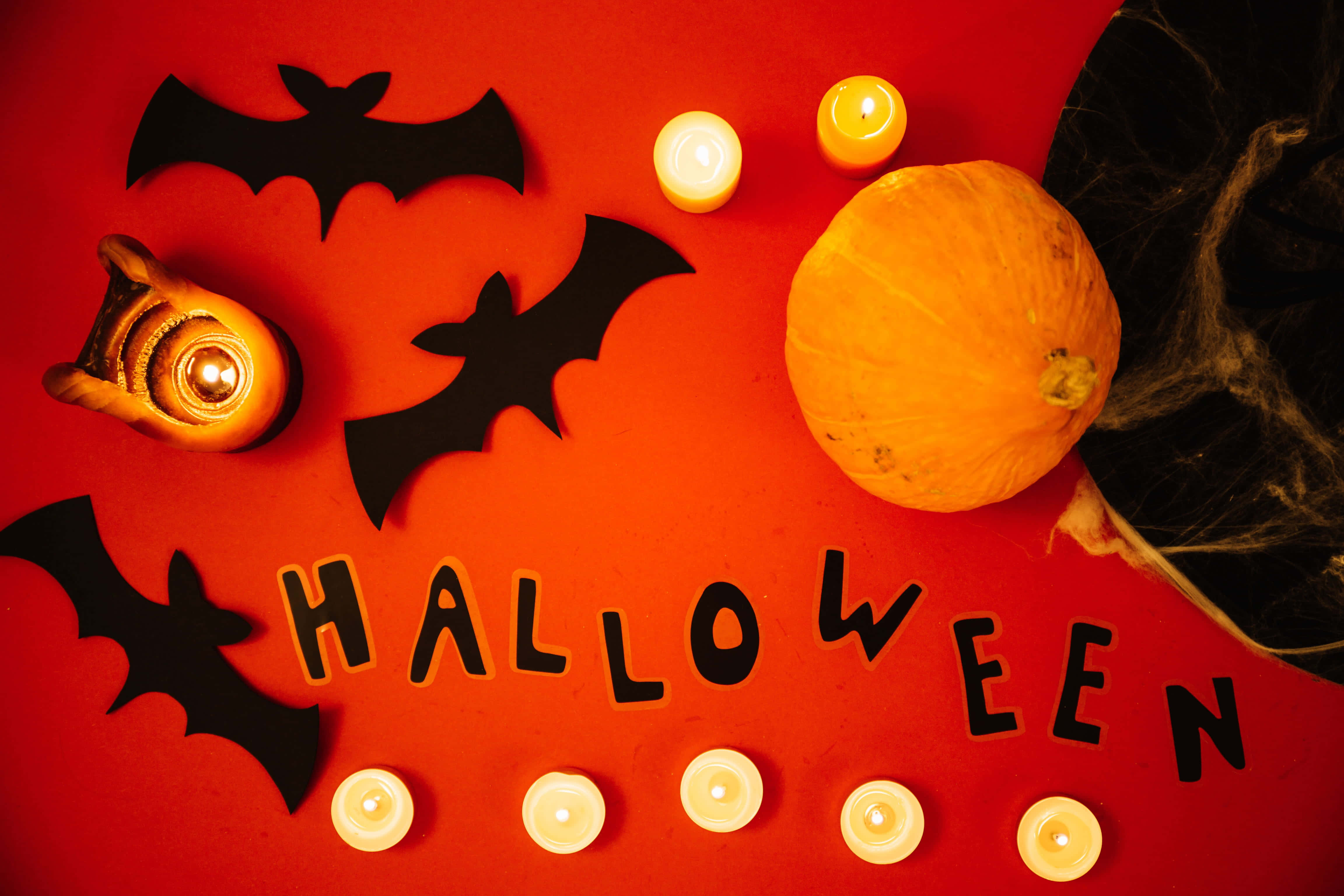 Halloween Candles 桌布 壁紙 — 一個節日萬聖節平鋪擺拍，有發光蠟燭、蝙蝠、南瓜和單詞Halloween，背景是鮮豔的紅色。