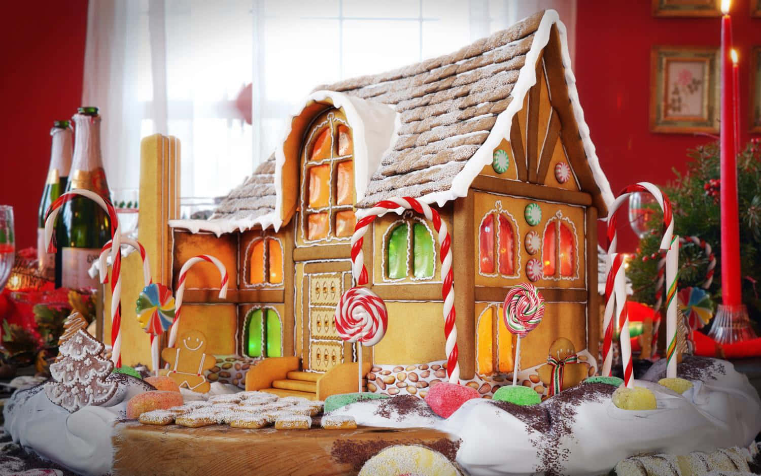 Gingerbread 桌布 壁紙 — 一個節日薑餅屋，裝飾有拐杖糖、棒棒糖和灑滿雪的屋頂，放置在木製底座上，周圍環繞著節日美食。