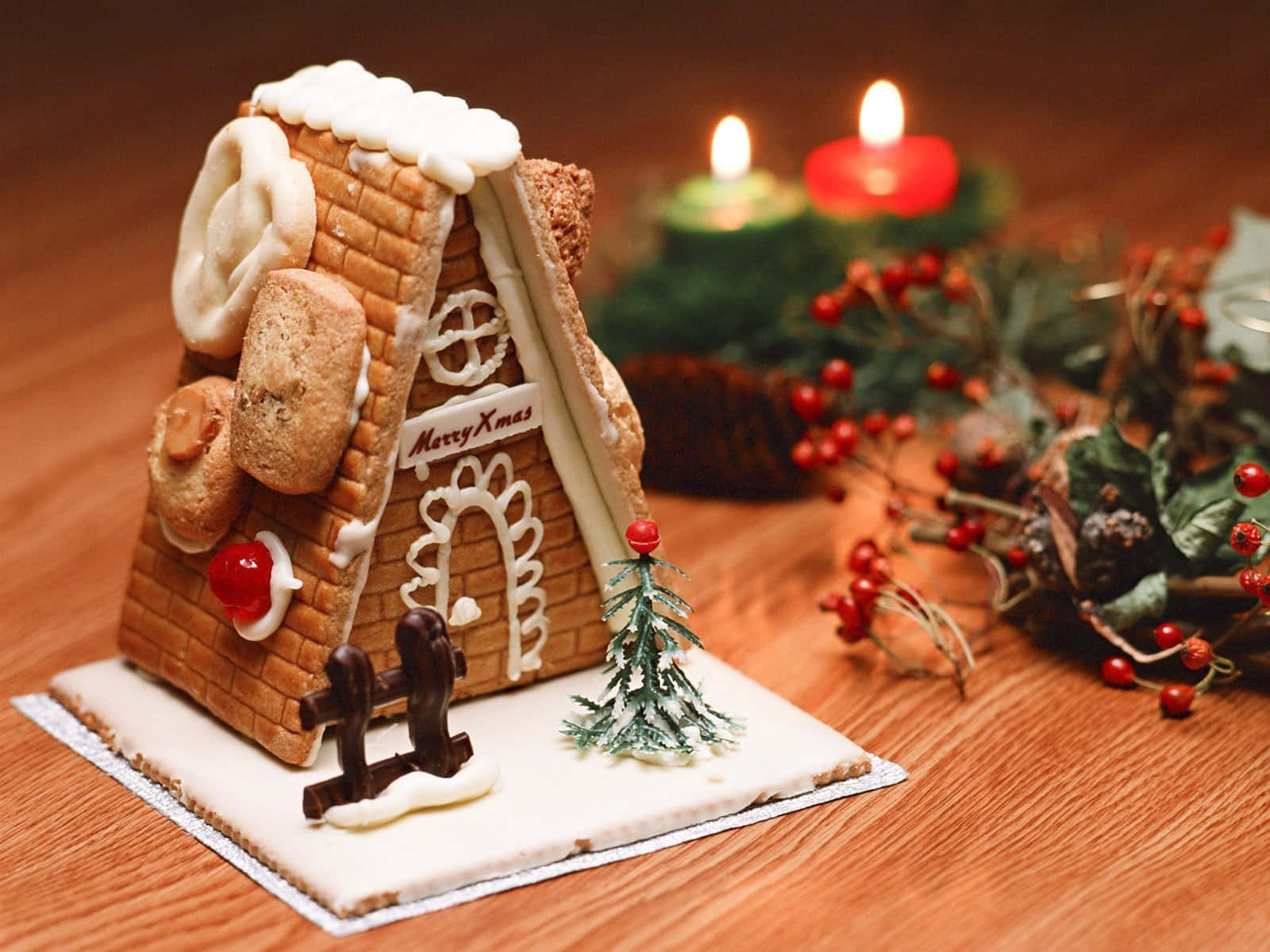 Gingerbread 桌布 壁紙 — 一個節日薑餅屋，飾有糖霜雪和糖果裝飾，放在木桌上，旁邊有發光的蠟燭和紅莓。