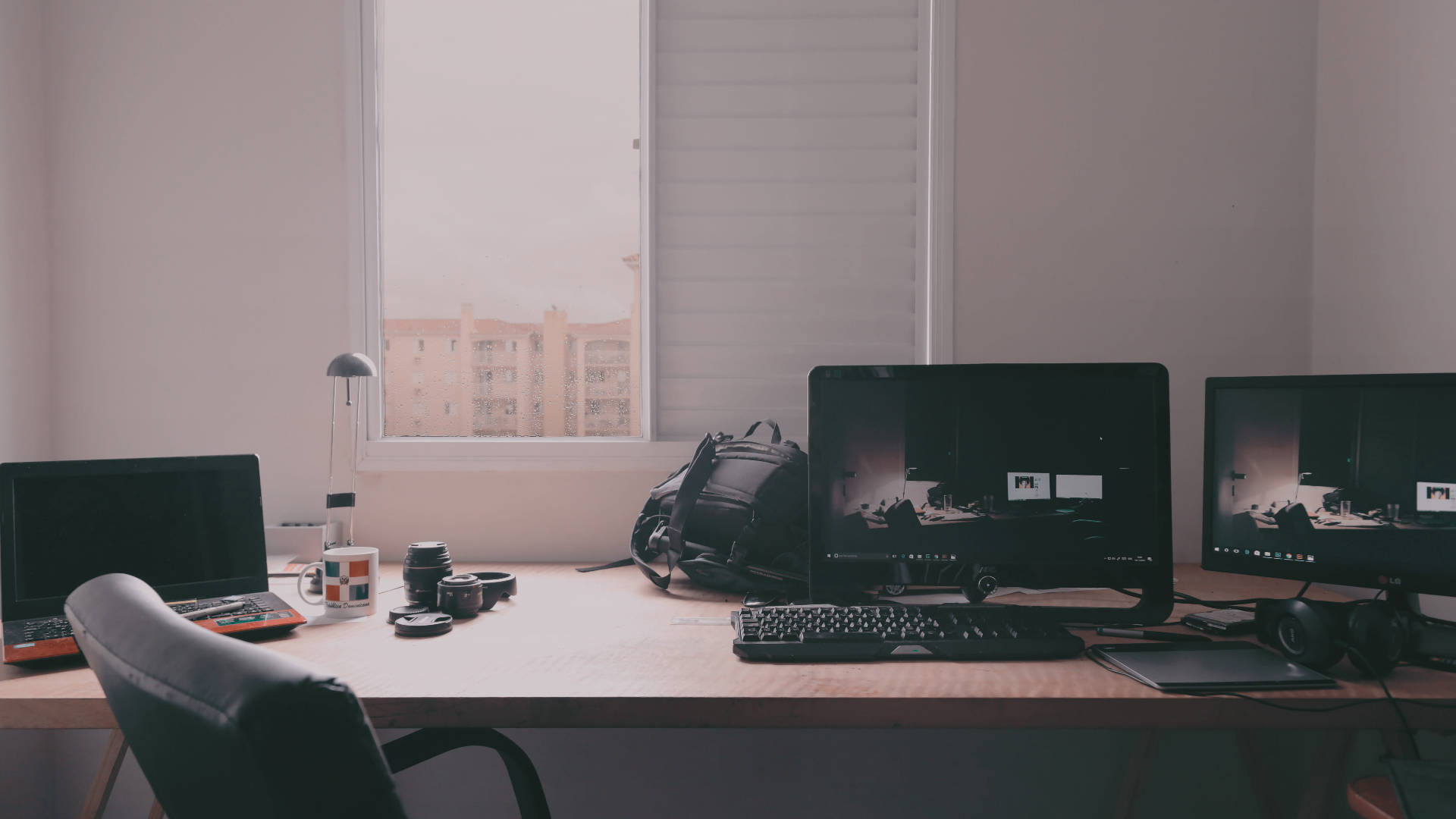 Working From Home 桌布 壁紙 — 一個簡約的家居辦公室書桌，配備雙顯示器、相機和手提電腦，沐浴在從窗戶透入的柔和自然光中，窗外可俯瞰城市建築。