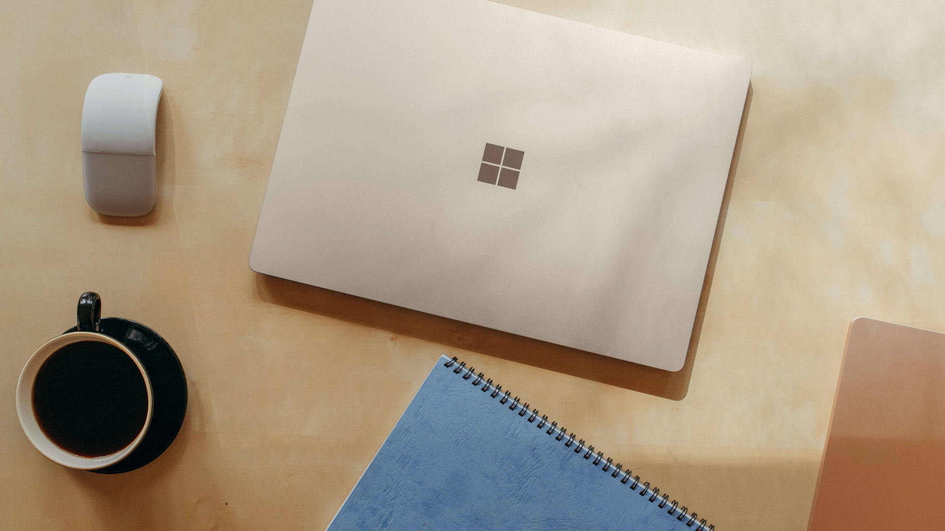 microsoft 桌布 壁紙 — 一個簡約的工作空間，在溫暖的木桌上放著一部關閉的銀色手提電腦、咖啡杯和筆記本。