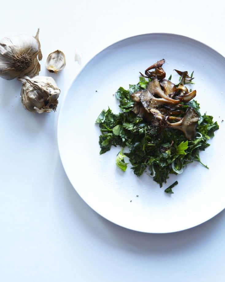 Black Garlic 桌布 壁紙 — 一個簡約的白色碟子上放著炒蘑菇，下面是鮮綠色的羽衣甘藍，旁邊還有整顆大蒜，放在潔白的表面上。