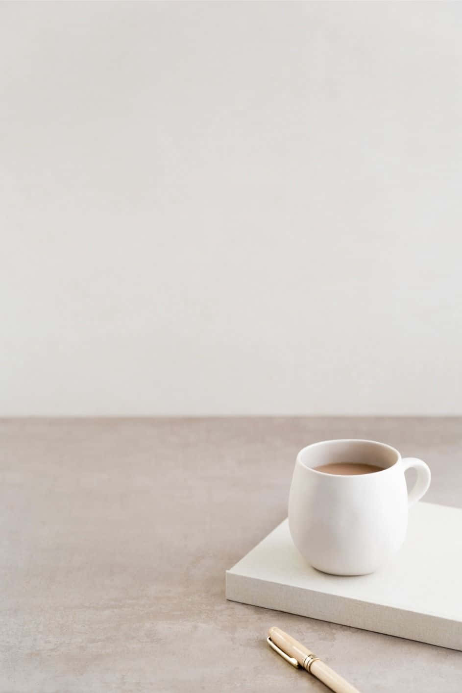 Minimalist Photography 桌布 壁紙 — 一個簡約的白色陶瓷杯裝滿咖啡，放在一本白色書本上，旁邊有一支木筆，置於中性色調的表面上。