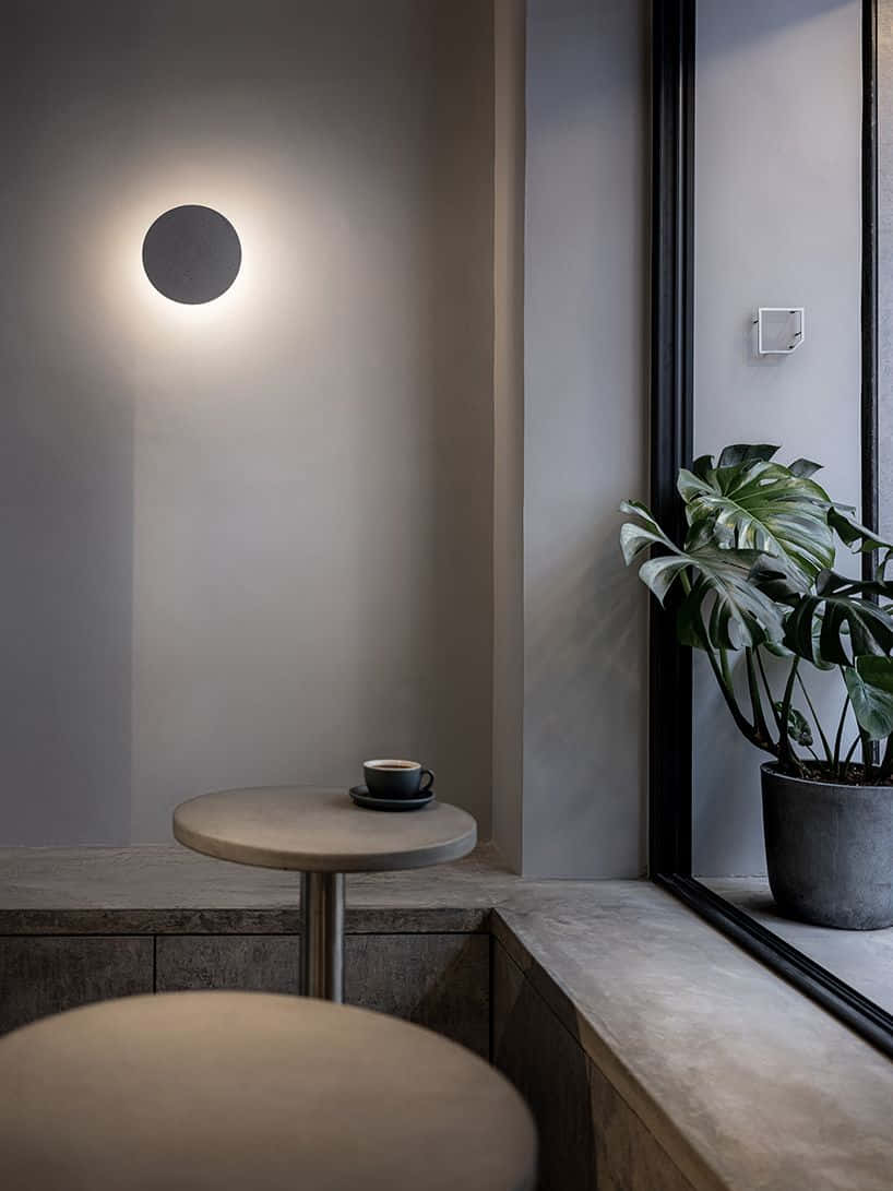 Minimalist Coffee 桌布 壁紙 — 一個簡約風格的咖啡店角落，圓桌上放著一杯咖啡，旁邊是混凝土花盆中的大型龜背竹，上方有圓形壁燈。