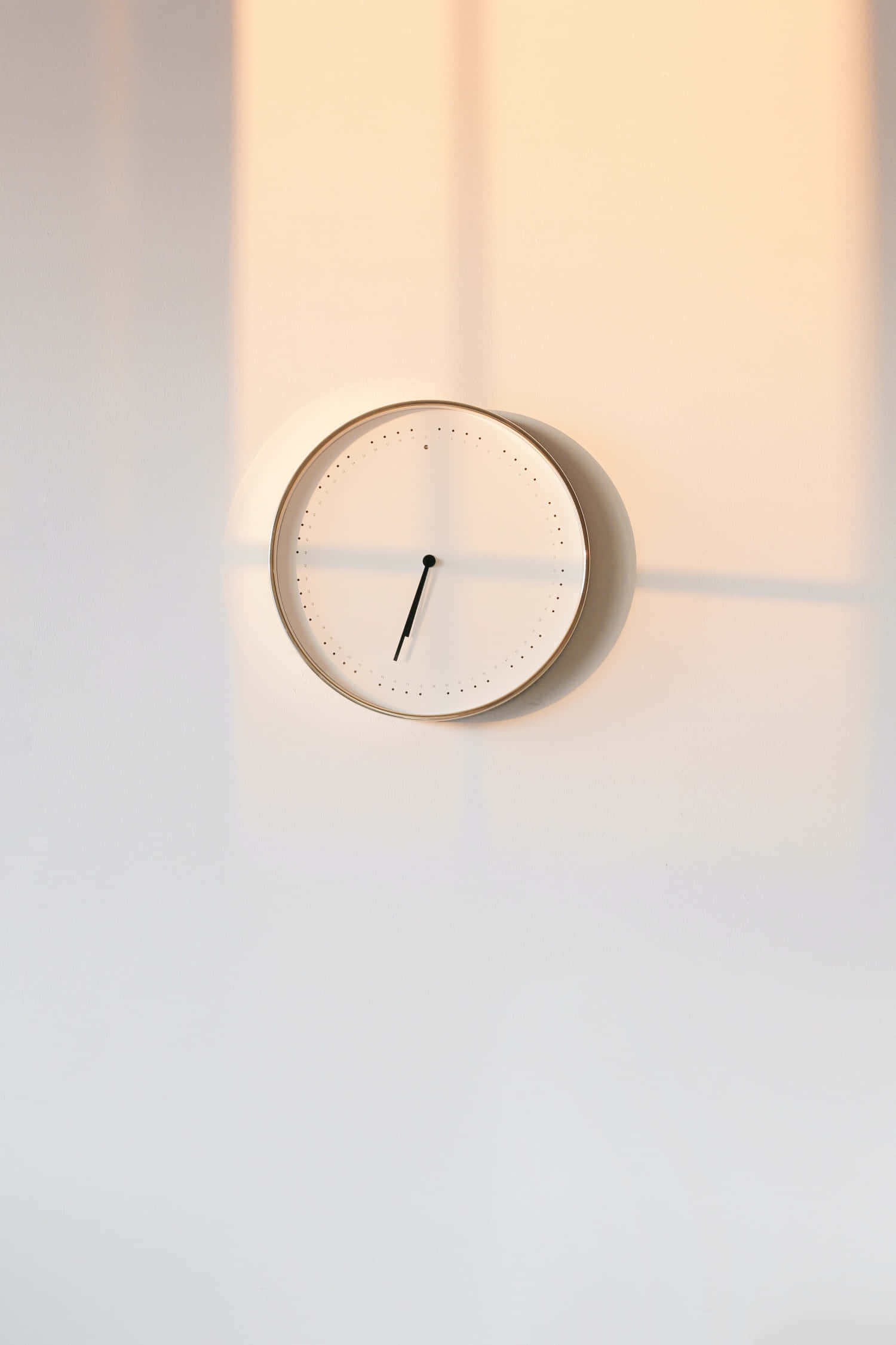 Minimalism 桌布 壁紙 — 一個簡約風格的掛牆時鐘，帶有金色邊框和黑色指針，在溫暖陽光照射下的白色牆壁上投下柔和陰影。