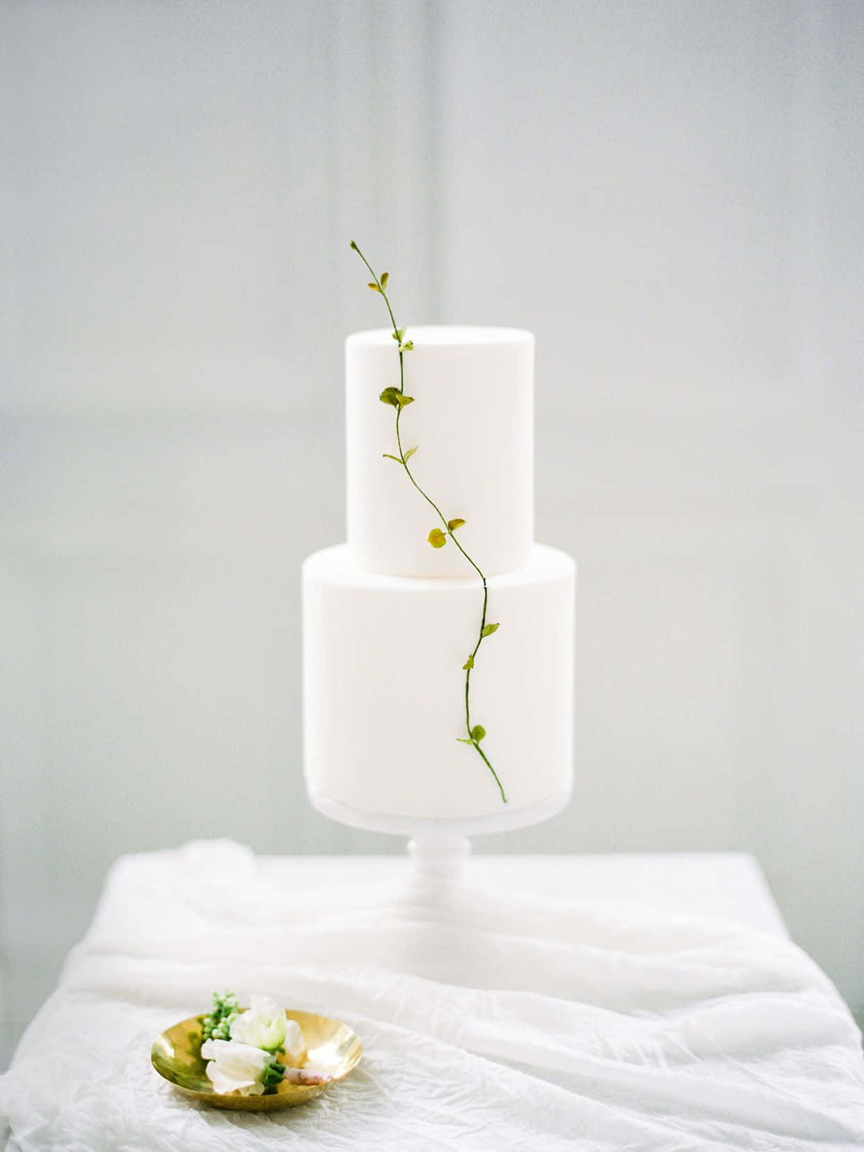 Cake Fondant 桌布 壁紙 — 一個簡約風格的白色兩層結婚蛋糕，飾以單條綠色藤蔓和小黃花，優雅地放在白色底座上，旁邊有一個金色盤子盛著白色花朵。