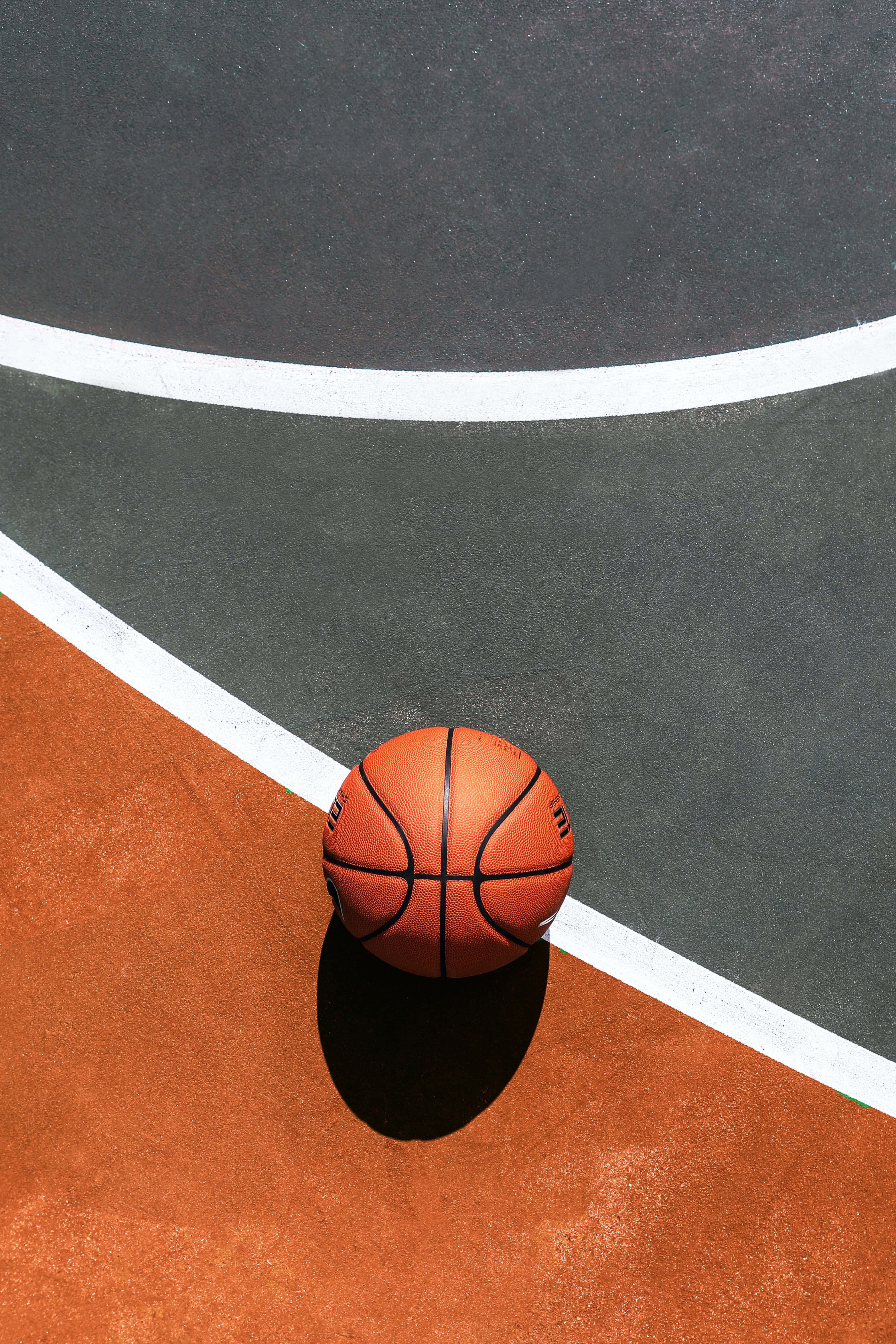 Minimalist Sports 桌布 壁紙 — 一個籃球放在球場的橙色和灰色表面上，表面有清晰的白色線條，在明亮的陽光下。