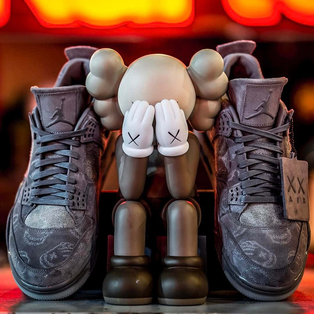 Kaws X Air Jordan 桌布 壁紙 — 一個米色KAWS公仔交叉雙手，坐在兩對灰色Jordan運動鞋之間，下方是發光的紅色霓虹燈。