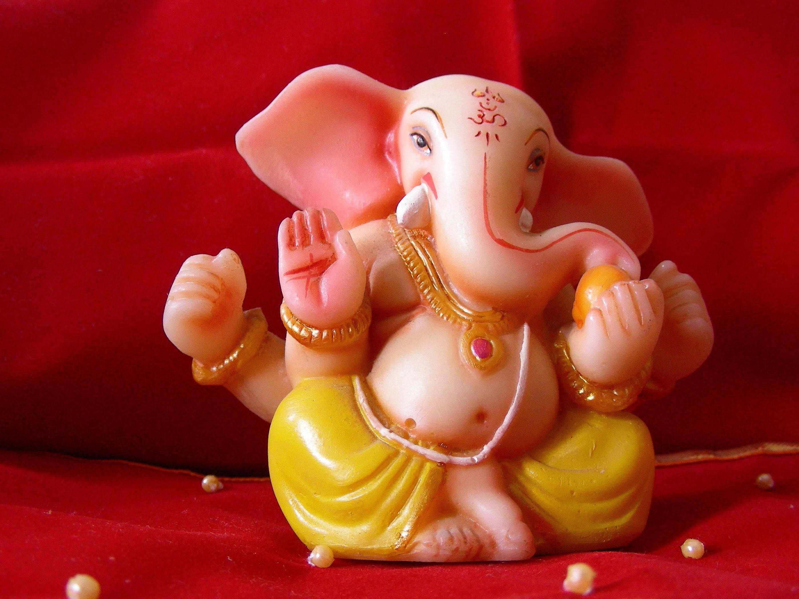 Hindu God 桌布 壁紙 — 一個粉紅色塑膠象頭神像，飾有金色珠寶和黃色腰布，坐在華麗的紅色天鵝絨表面上，周圍散落著珍珠。