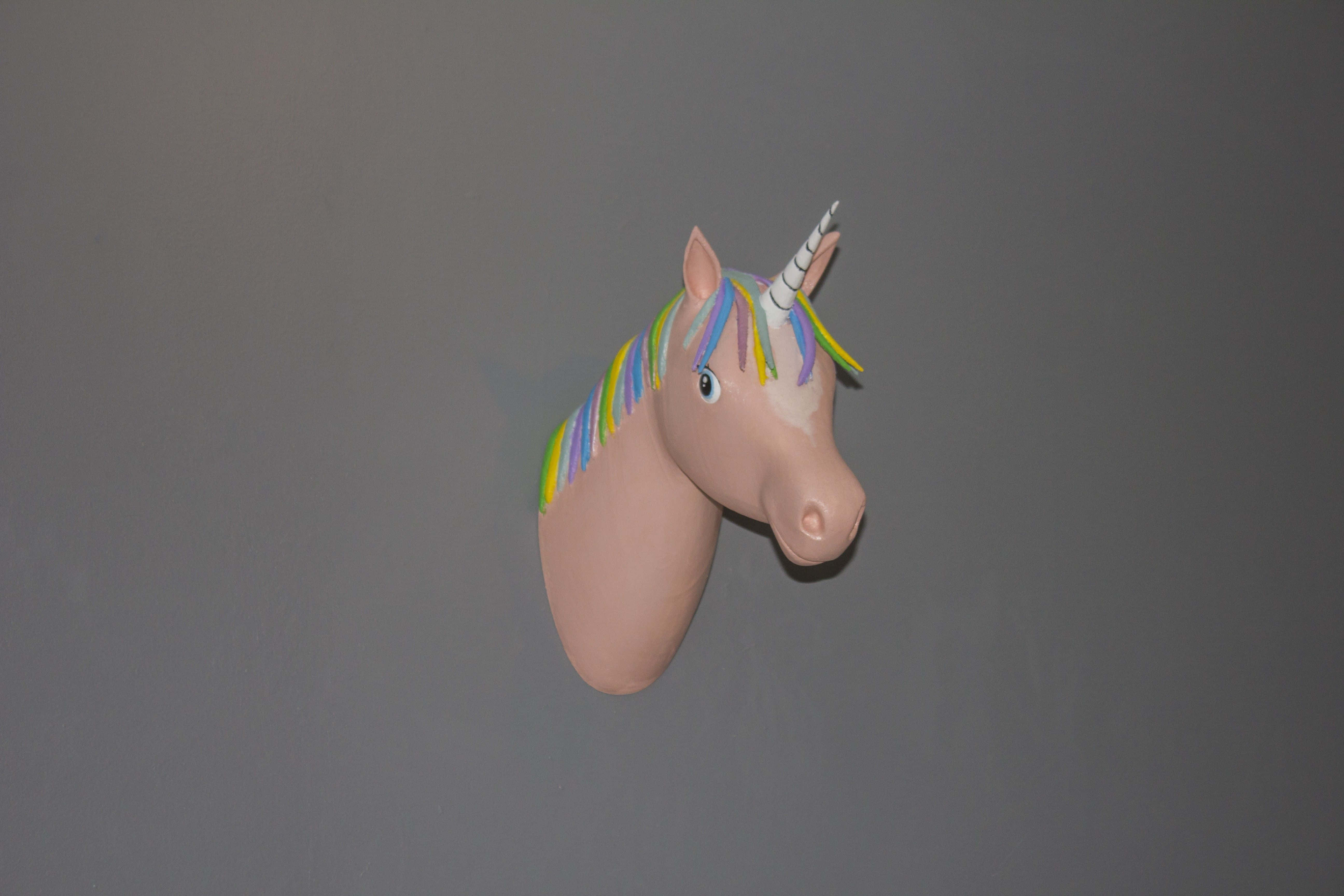 3d Unicorn 桌布 壁紙 — 一個粉紅色獨角獸頭部裝飾，帶有彩虹鬃毛和角，掛在純灰色牆上。