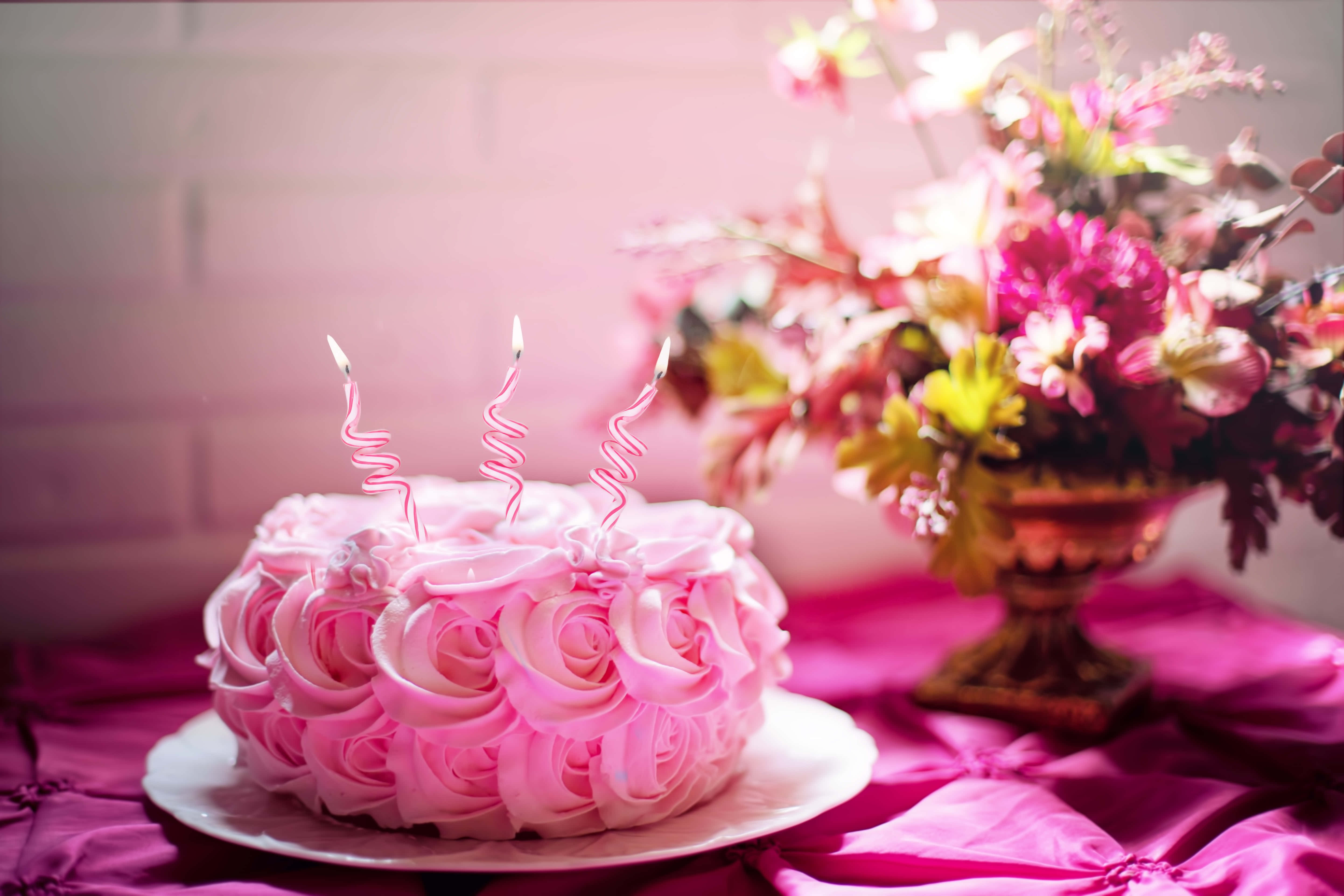 Floral Cake 桌布 壁紙 — 一個粉紅色玫瑰形狀的生日蛋糕，上面有三根點燃的螺旋蠟燭，旁邊是一個充滿活力的插花，放在洋紅色緞布上。