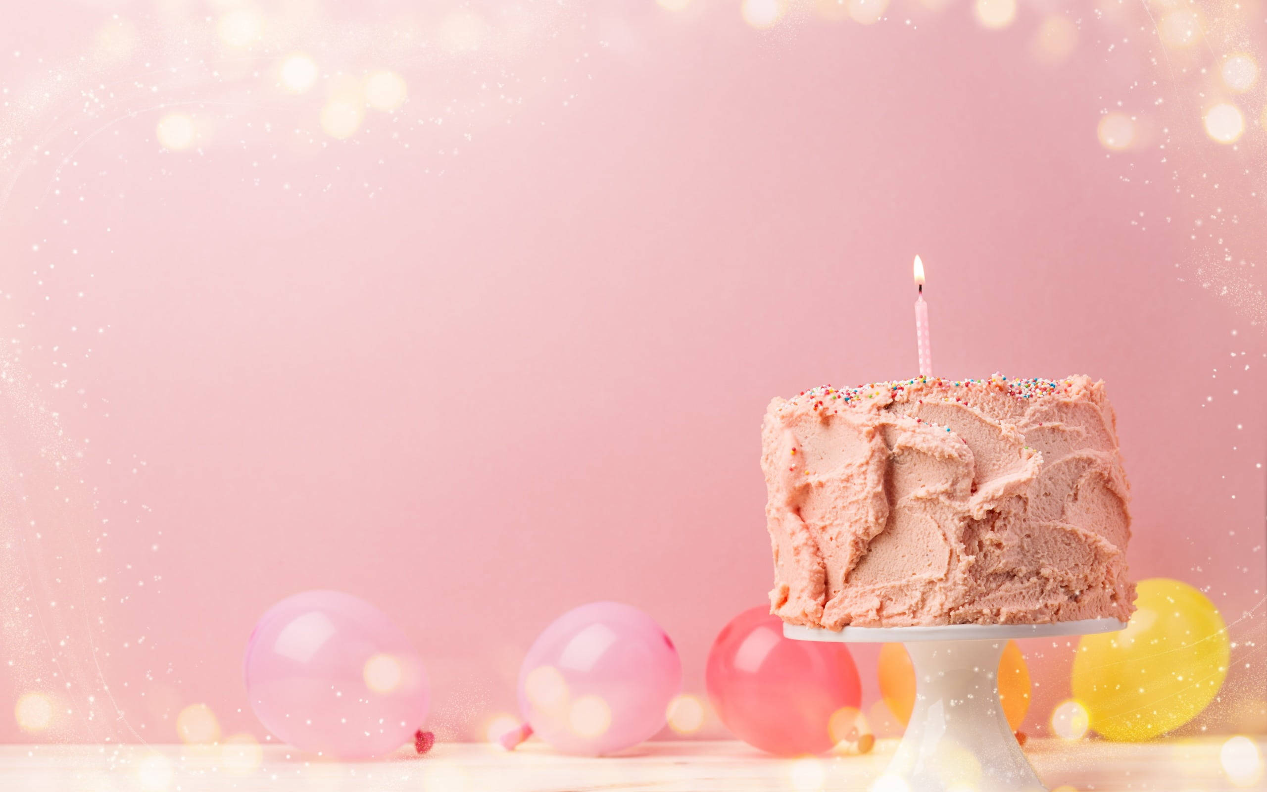 生日蛋糕 桌布 壁紙 — 一個粉紅色糖霜生日蛋糕，上面插著一支蠟燭，放在一個被彩色氣球環繞的架子上，背景是柔和的粉紅色散景。