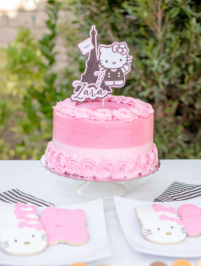 Hello Kitty Party 桌布 壁紙 — 一個粉紅色糖霜生日蛋糕，上面放著Hello Kitty和艾菲爾鐵塔裝飾，放在架子上，旁邊有主題曲奇。