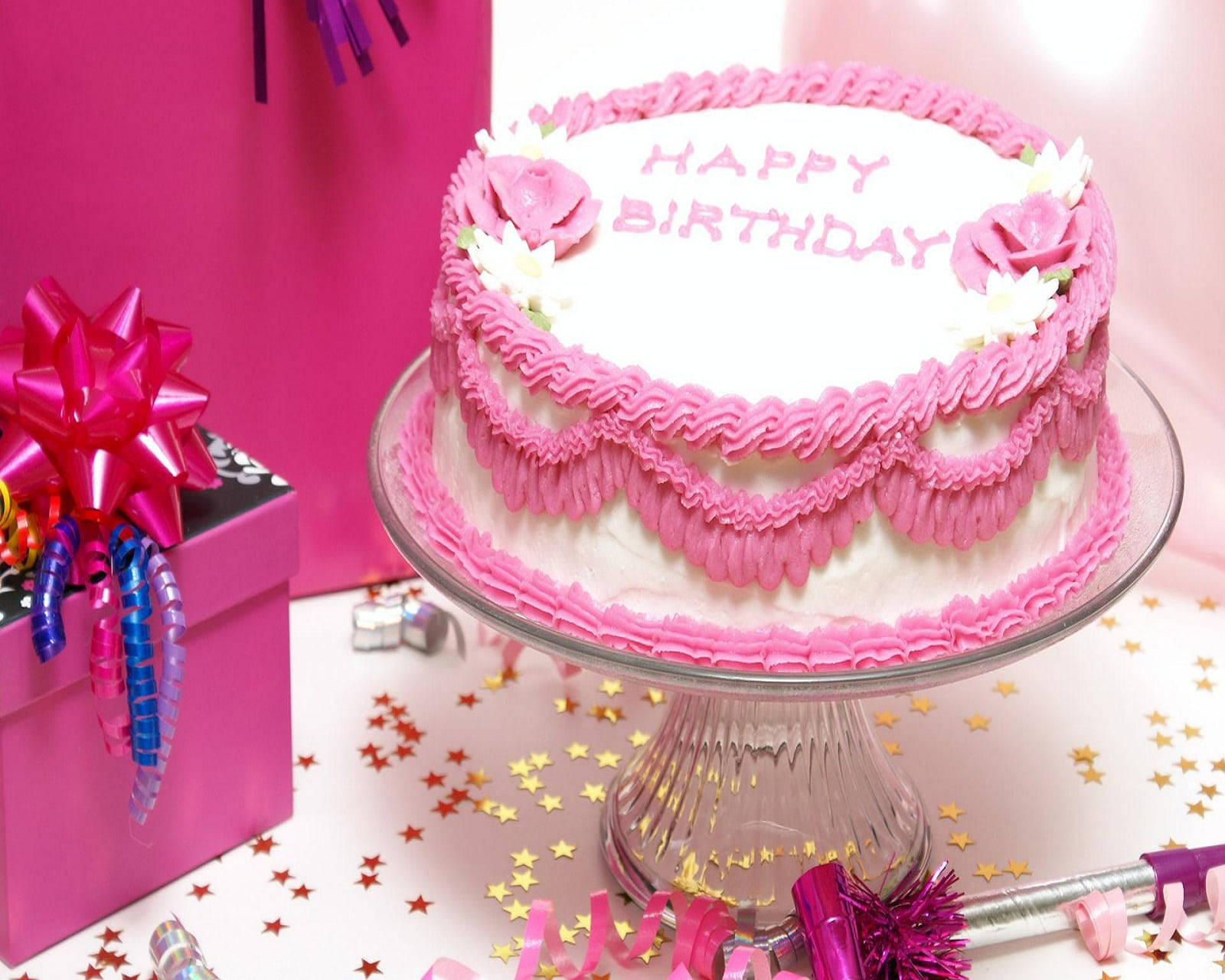 Birthday 桌布 壁紙 — 一個粉紅色糖霜生日蛋糕，帶有褶邊糖霜和花朵，放在玻璃架上，旁邊有一個禮物盒和金色星星紙屑。