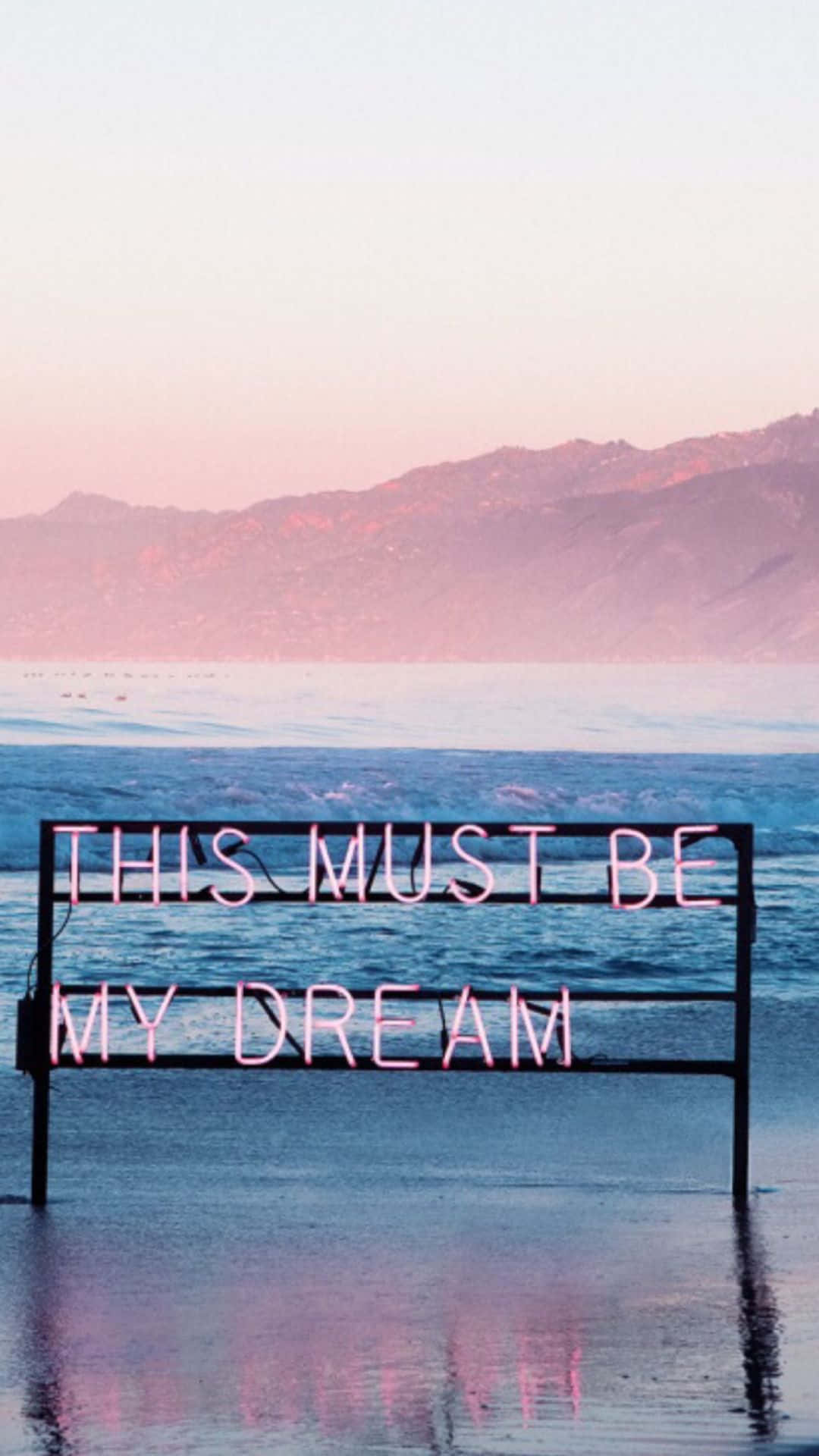 美學手機GIF 桌布 壁紙 — 一個粉紅色霓虹燈牌，上面寫著「THIS MUST BE MY DREAM」，矗立在日落時分的淺海水中，背景有山脈。