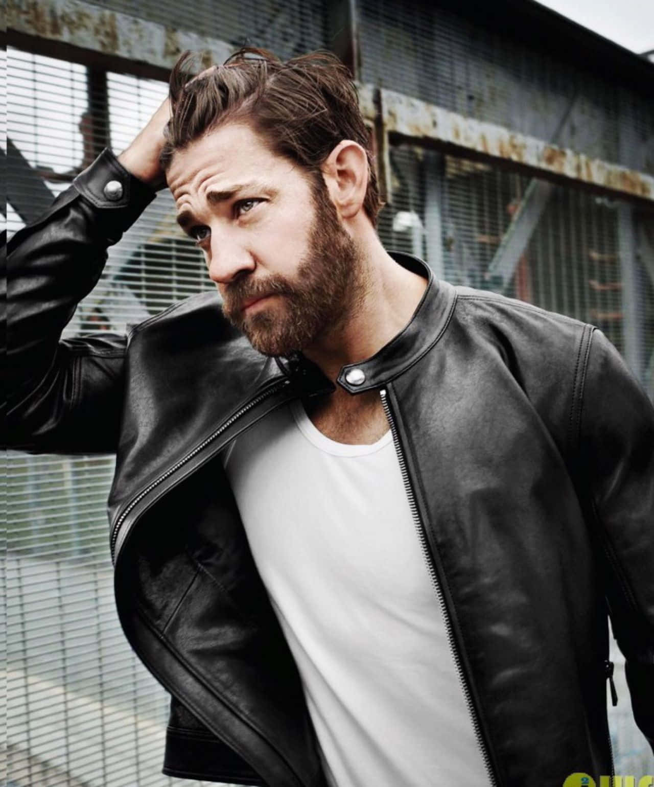 john krasinski 桌布 壁紙 — 一個粗獷留鬚的男人穿著黑色皮褸，在生鏽的工業圍欄背景下用手撥弄頭髮。