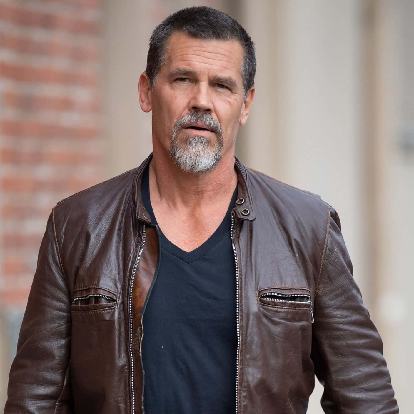 Josh Brolin 桌布 壁紙 — 一個粗獷的男人留著灰白鬍鬚，身穿棕色皮褸內搭黑色T恤，背景係磚牆。
