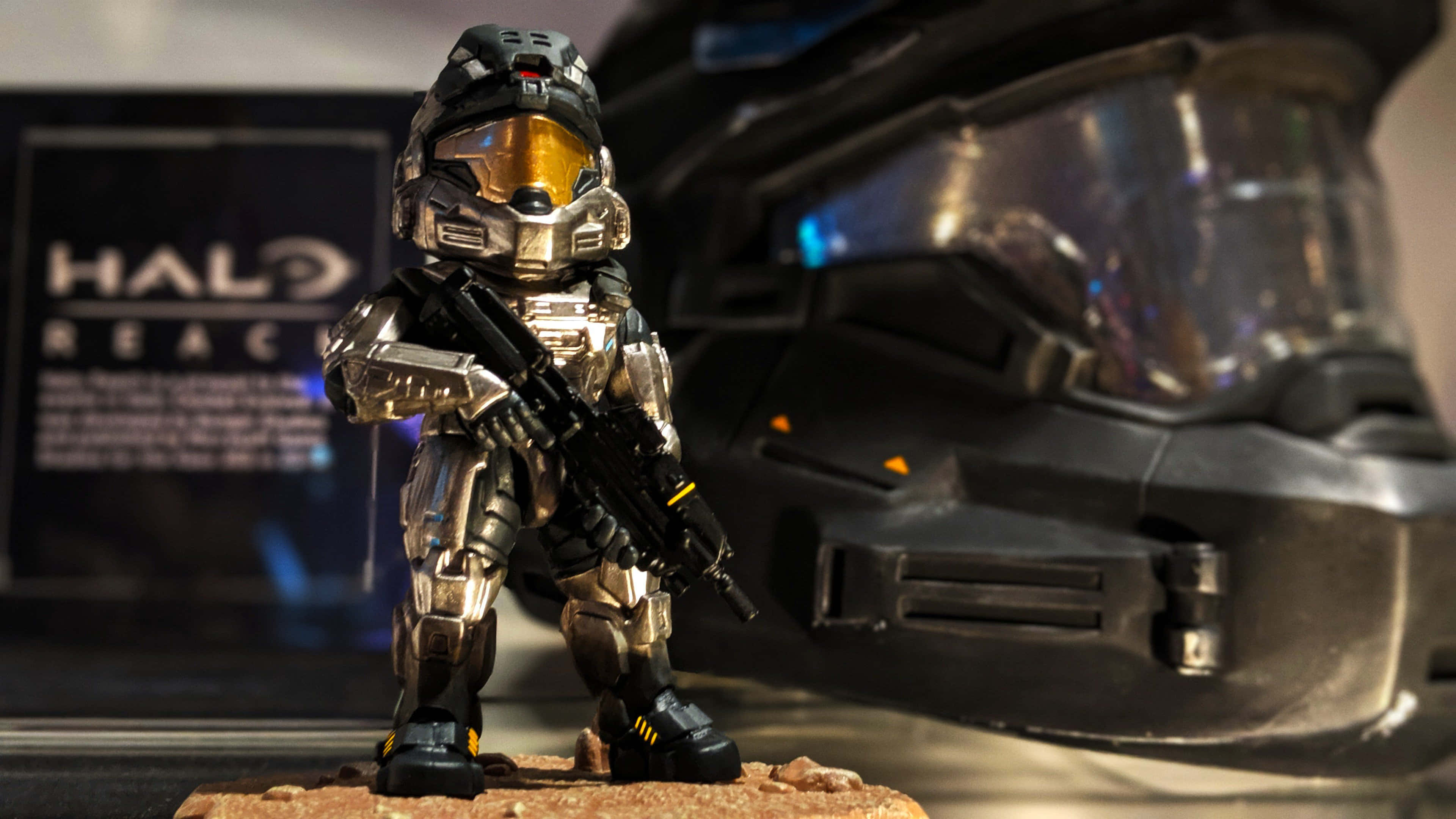 Noble Six 桌布 壁紙 — 一個精細的Halo Reach斯巴達人偶手持步槍，站在一個大頭盔和遊戲盒旁邊，置於收藏家展示櫃中。