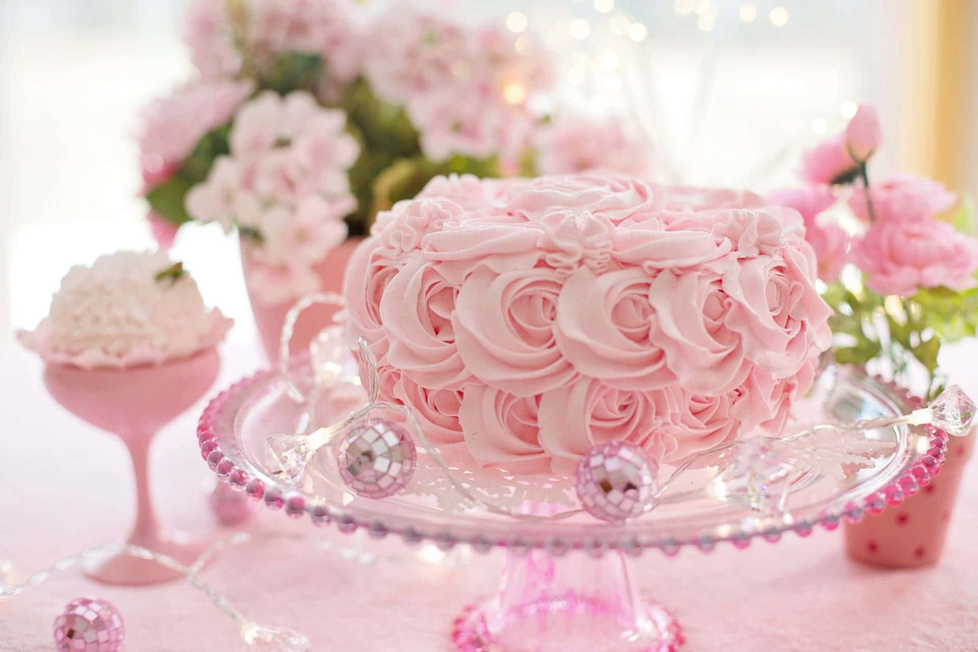 Cake Fondant 桌布 壁紙 — 一個精緻的粉紅色蛋糕，飾有玫瑰形狀的糖霜，放在玻璃架上，周圍環繞著粉紅色花朵和迪斯科球。