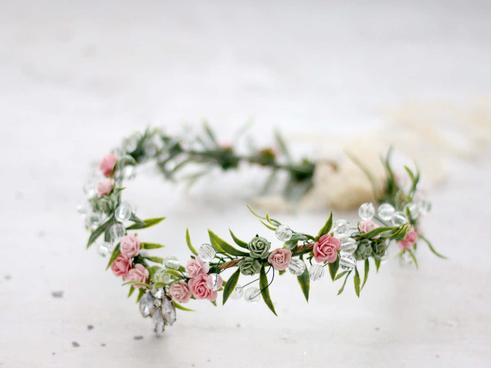 Floral Crown 桌布 壁紙 — 一個精緻的花冠，點綴著粉紅玫瑰、綠葉和透明水晶，輕輕地放在有紋理的白色表面上。