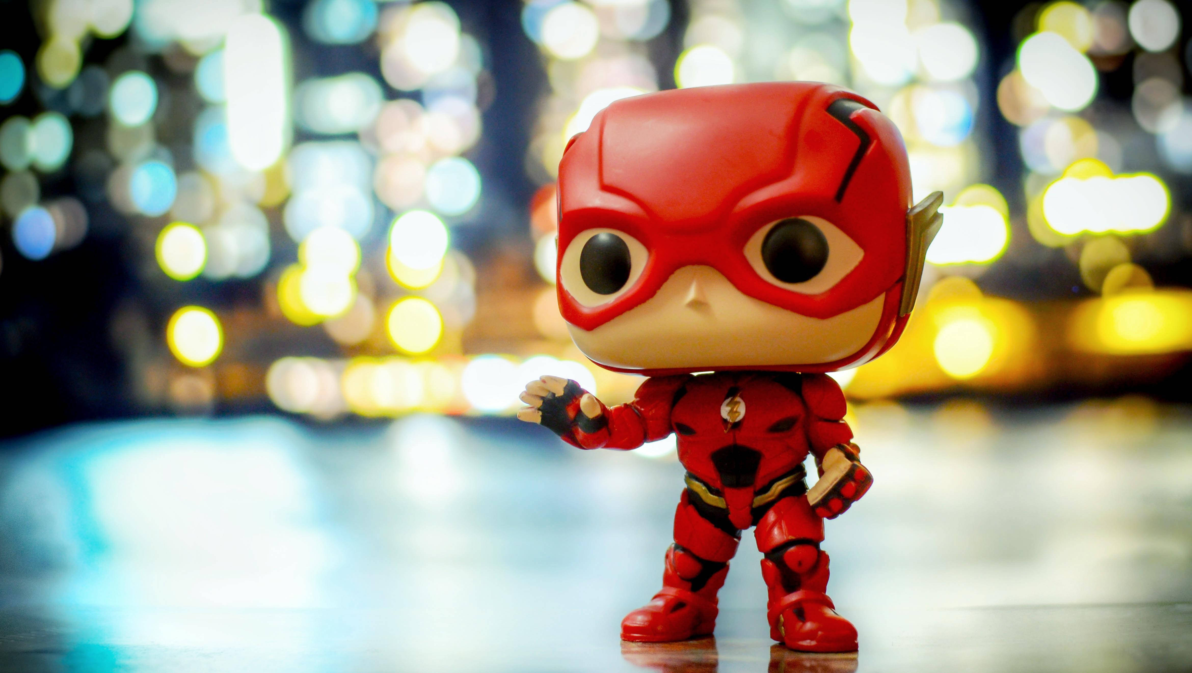 Funko Pop Flash 壁紙 — 一個紅色Flash Funko Pop公仔自信地站在反光表面上，背景是充滿活力的散景城市夜景。