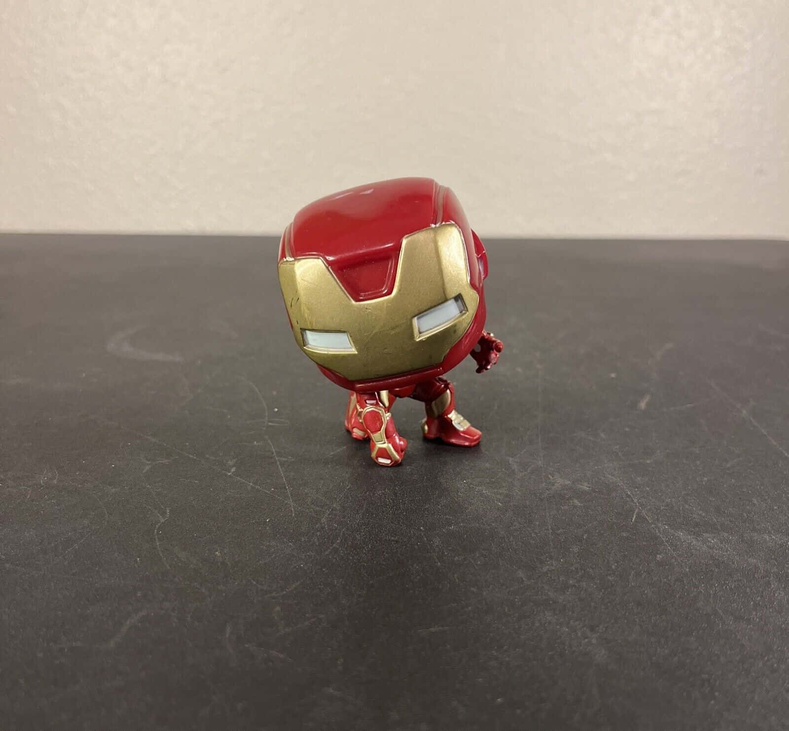 Iron Man Bobbleheads 桌布 壁紙 — 一個紅色和金色的鐵甲奇俠 Funko Pop 人偶，站立在一個有刮痕的深灰色表面上，背景是純色的米白色牆壁。