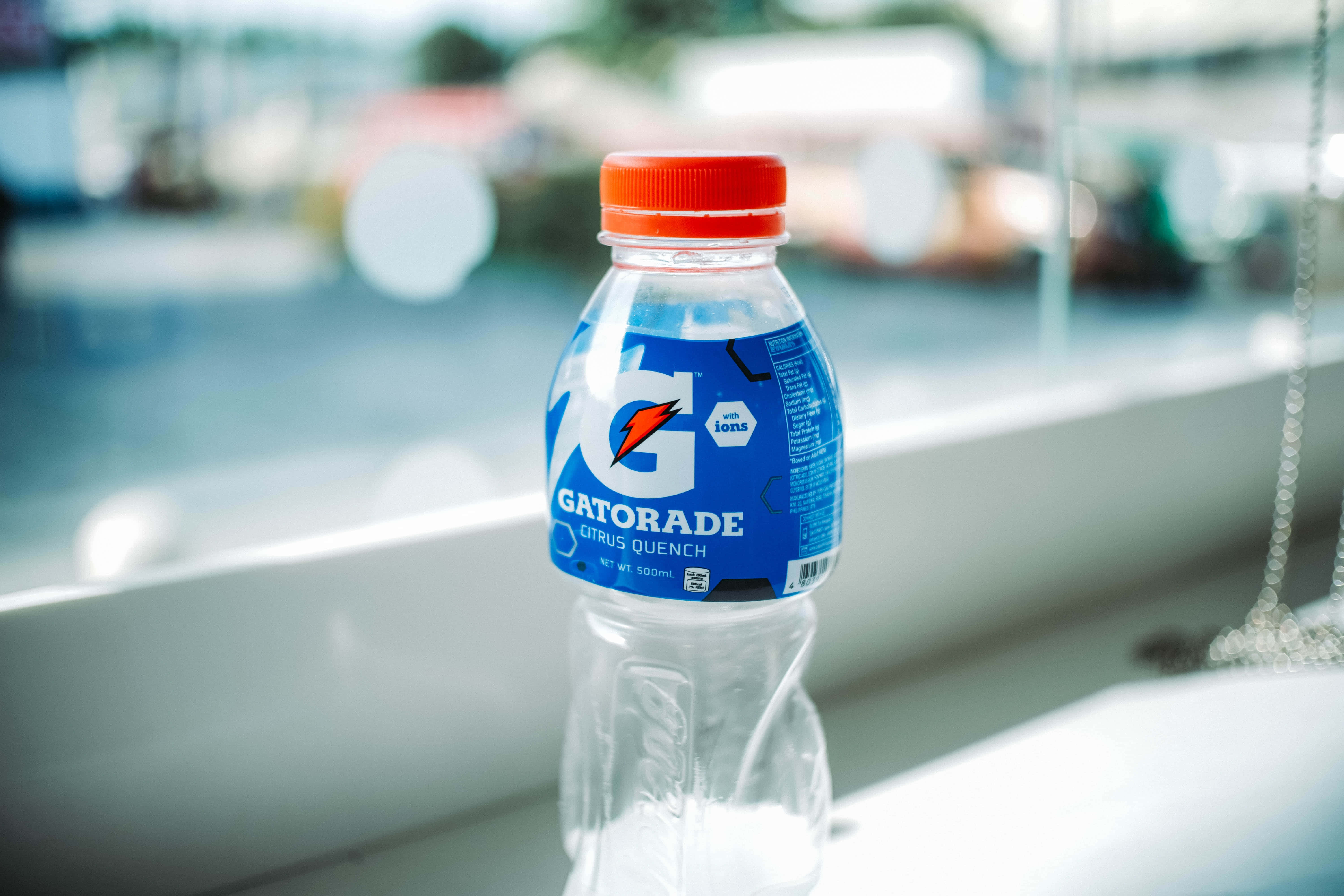 Hydration 桌布 壁紙 — 一個紅色蓋的透明Gatorade Citrus Quench樽放在白色窗台上，背景是模糊的戶外景色。