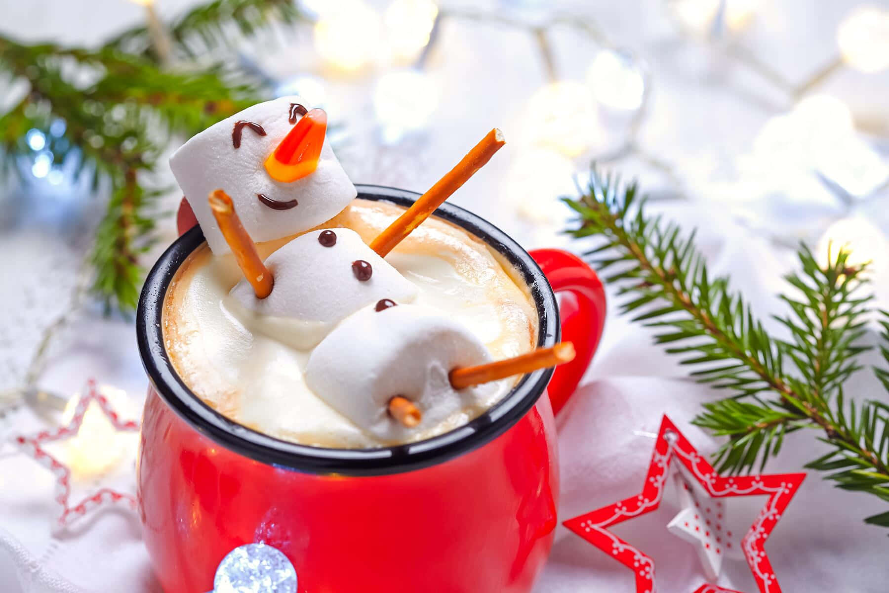 Winter Drinks 桌布 壁紙 — 一個紅色馬克杯，裝滿了奶油般的熱巧克力，上面放著一個用棉花糖、椒鹽脆餅棒和巧克力片做成的微笑棉花糖雪人。