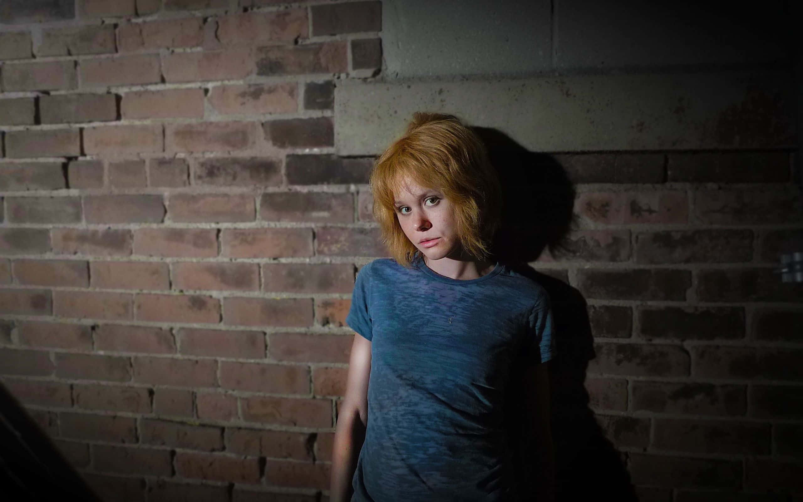 Alison Pill 桌布 壁紙 — 一個紅髮的年輕人靠在一面昏暗的磚牆上，在憂鬱的城市場景中投下長長的影子。