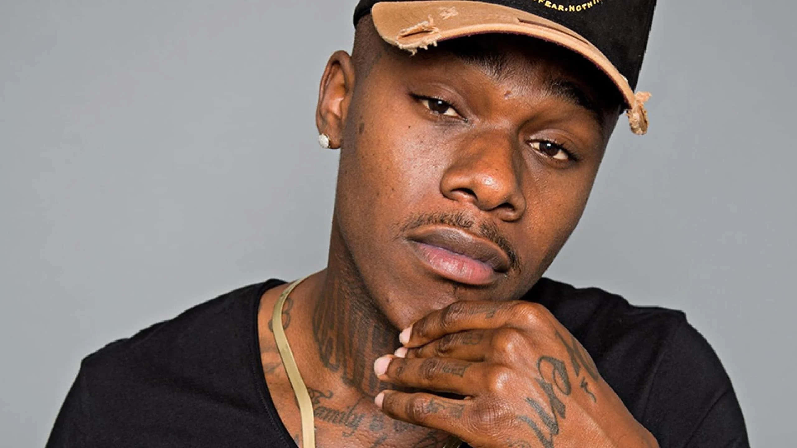 Dababy 桌布 壁紙 — 一個紋身男子的特寫肖像，戴著黑色帽子和襯衫，手托下巴，在純灰色背景上專注地凝視。