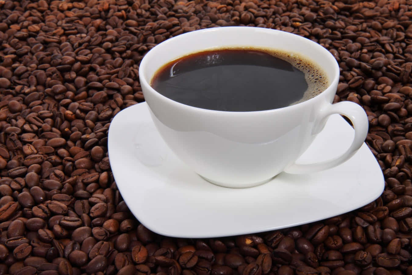 Coffee Cup 桌布 壁紙 — 一個純白色的咖啡杯盛載著深色咖啡，放在碟子上，置於烘焙咖啡豆之上。