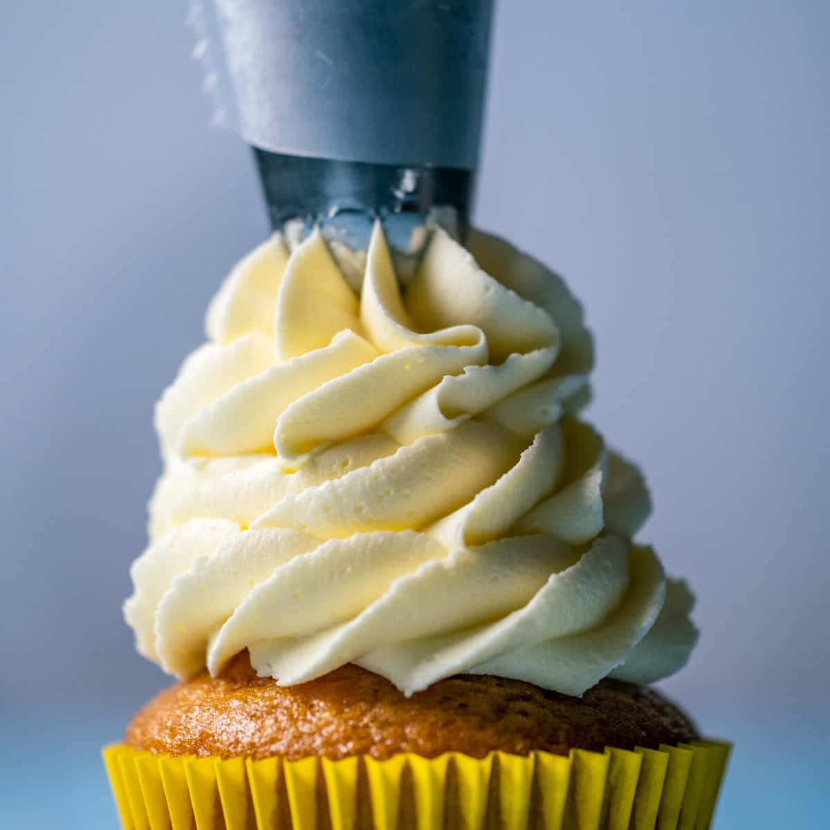 Buttercream 桌布 壁紙 — 一個紙杯蛋糕在黃色襯紙中，正在被擠上奶油雲呢拿糖霜，背景是柔和藍色的特寫。