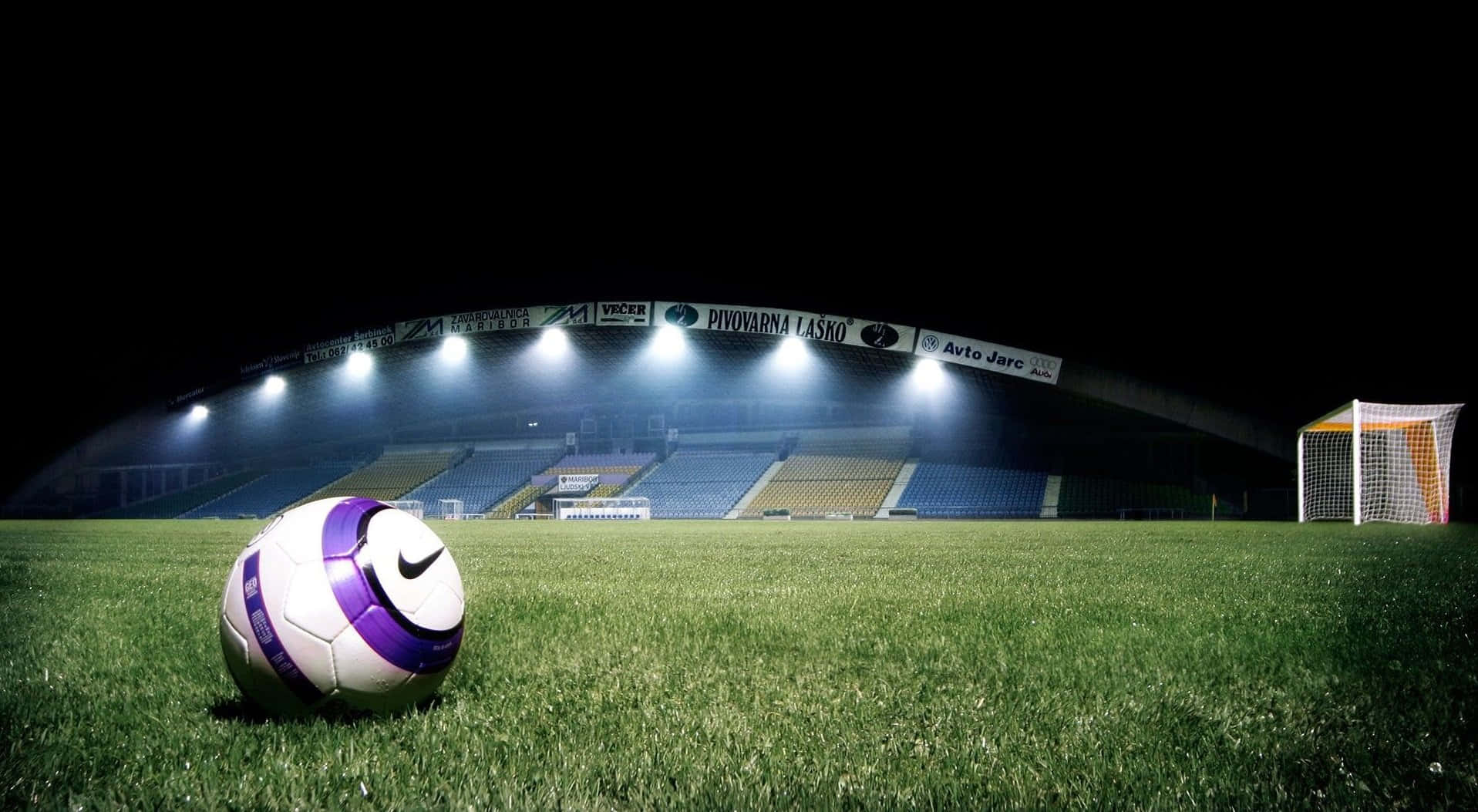 3d Sports 桌布 壁紙 — 一個紫色和白色的足球在夜間明亮的球場燈光下放在充滿活力的綠色草地上，背景是空蕩蕩的看台。