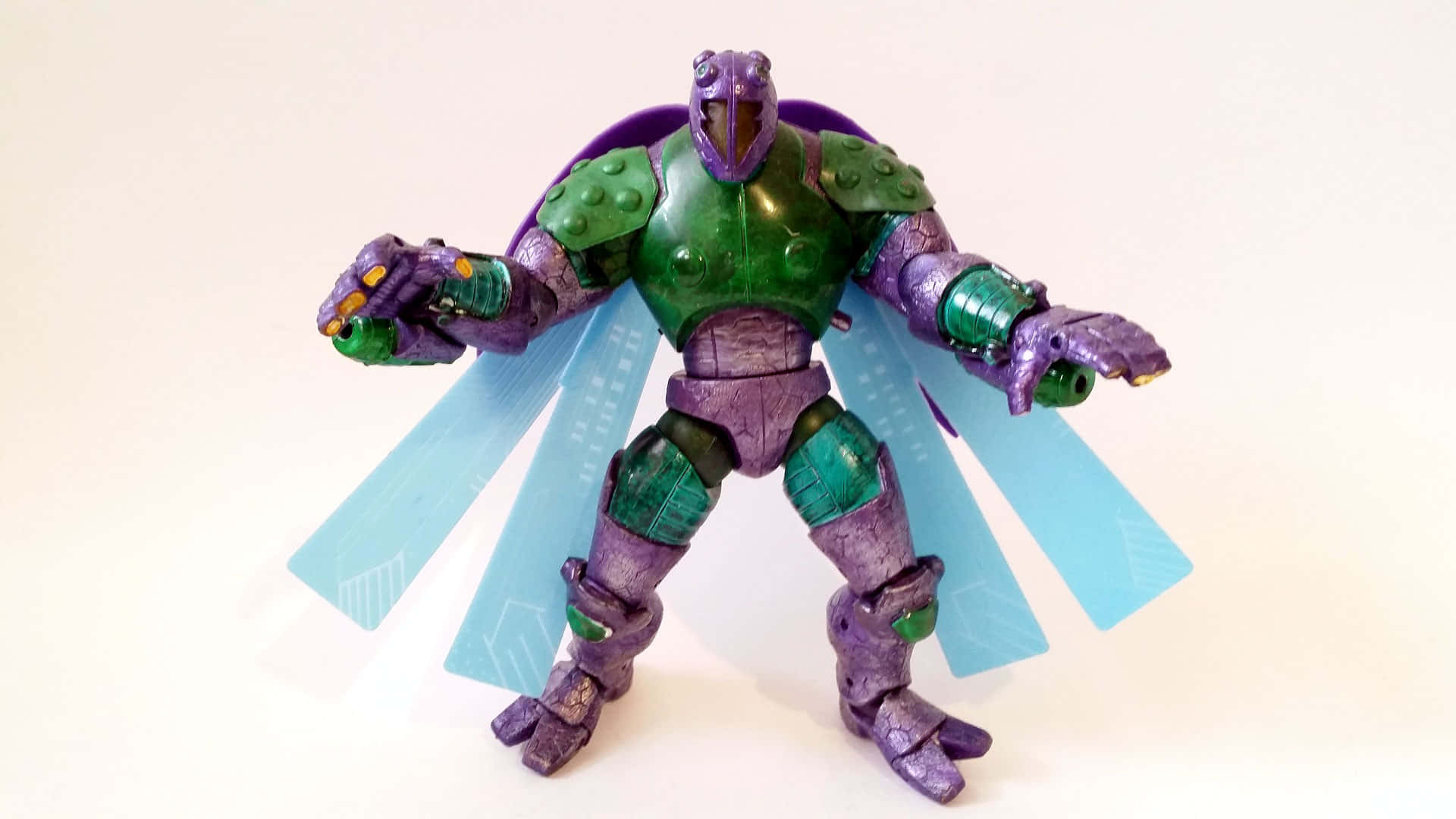 The Beetle 桌布 壁紙 — 一個紫色和綠色的裝甲動作人偶，披著藍色斗篷，在純白色背景前擺出動態姿勢。