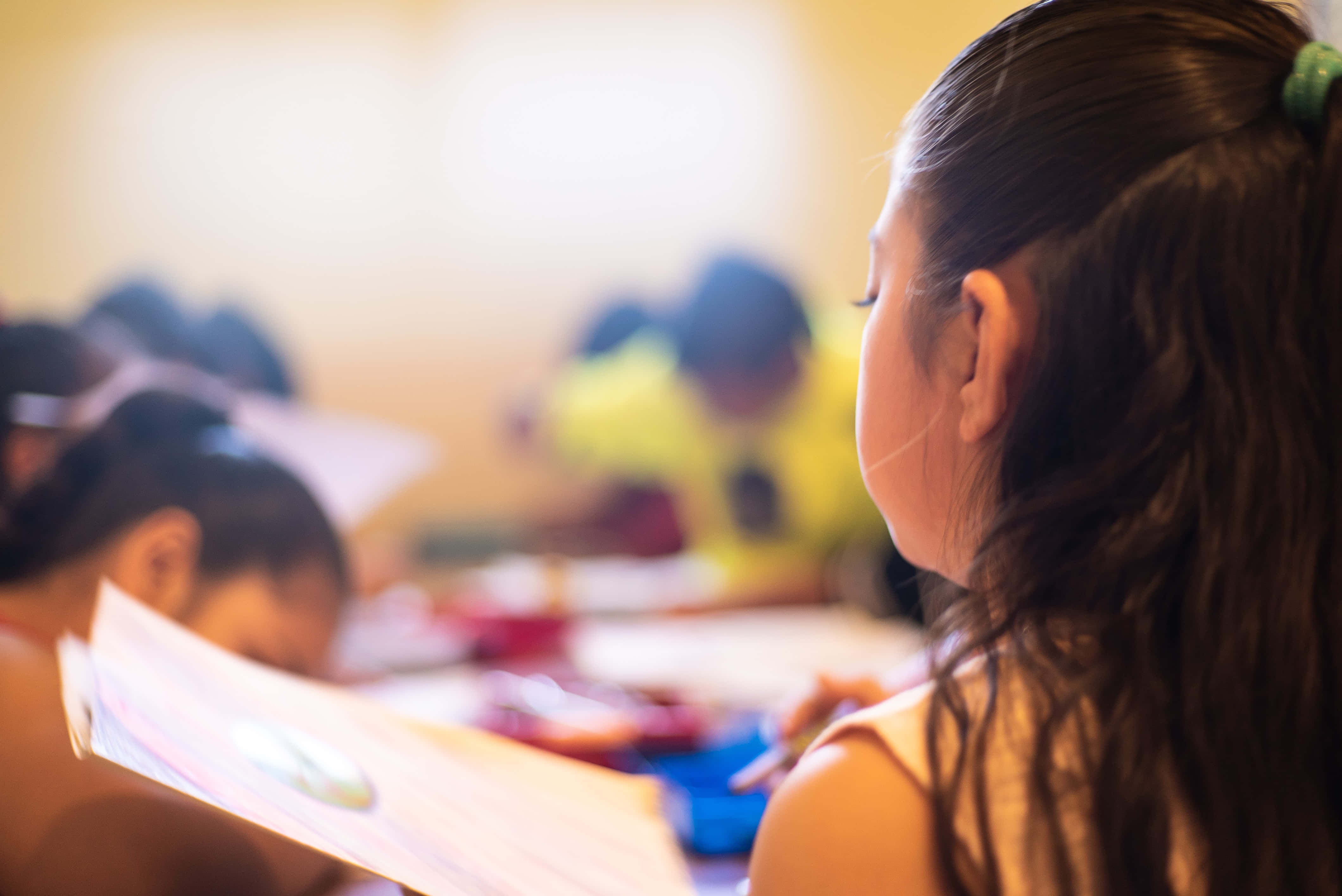 溫習 桌布 壁紙 — 一個紮著馬尾的黑髮女孩在溫暖光線的課室裡與其他孩子一起專注地畫畫。