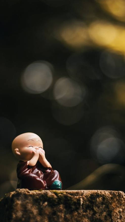 Buddha 桌布 壁紙 — 一個細小微笑嘅和尚公仔平靜咁坐喺粗糙嘅石頭上，後面有柔和嘅金色散景燈光閃爍。