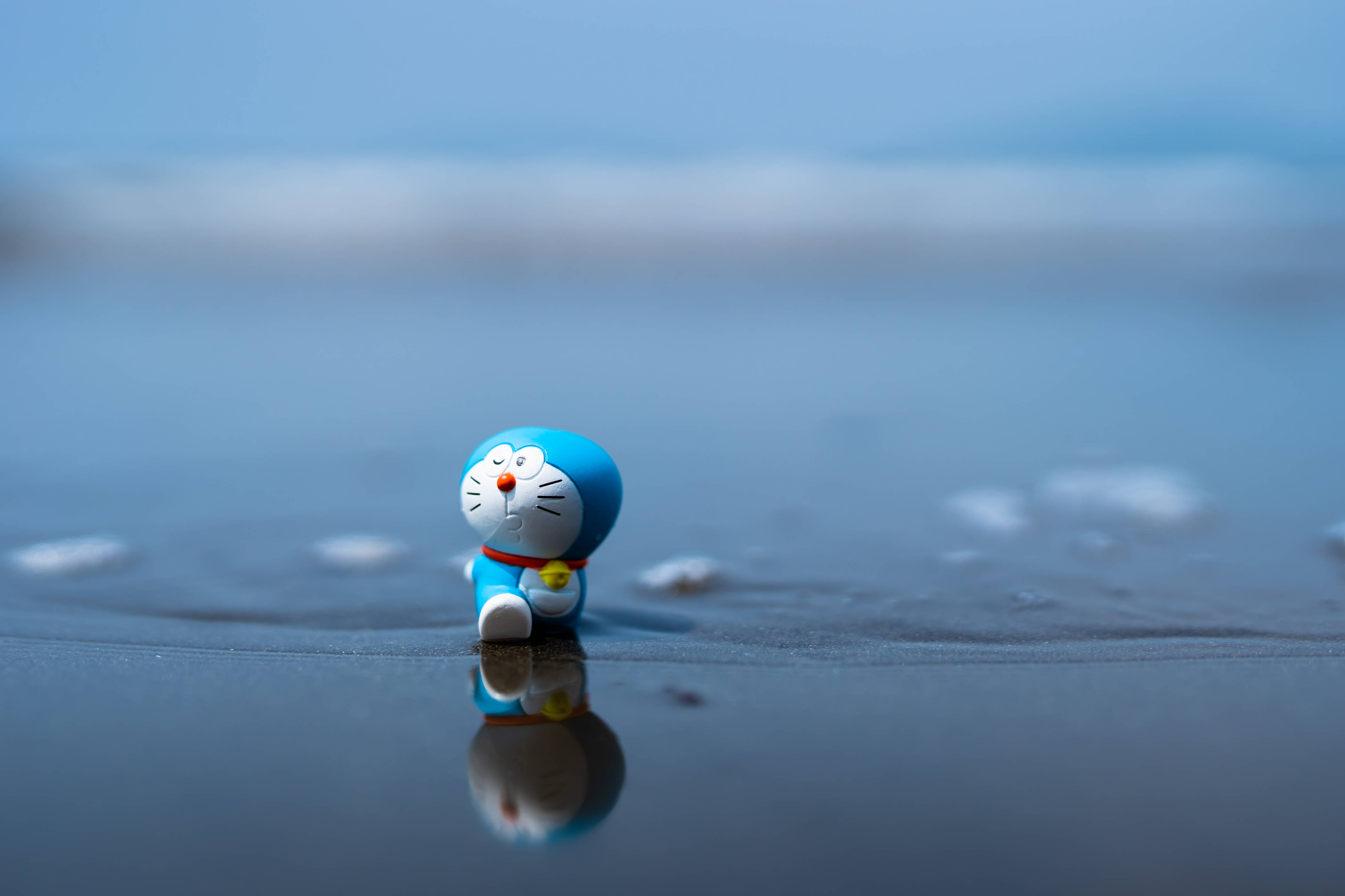 Doraemon 桌布 壁紙 — 一個細小的藍色多啦A夢公仔獨自坐在濕沙上，背景是模糊的海洋，並有清晰的倒影。
