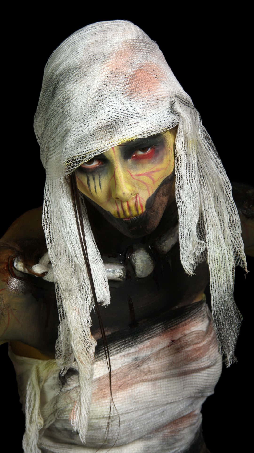 Mummy Costumes 桌布 壁紙 — 一個細節令人毛骨悚然的木乃伊，臉上塗著龜裂的黃色顏料，紅色的眼睛在黑色背景下凝視著。