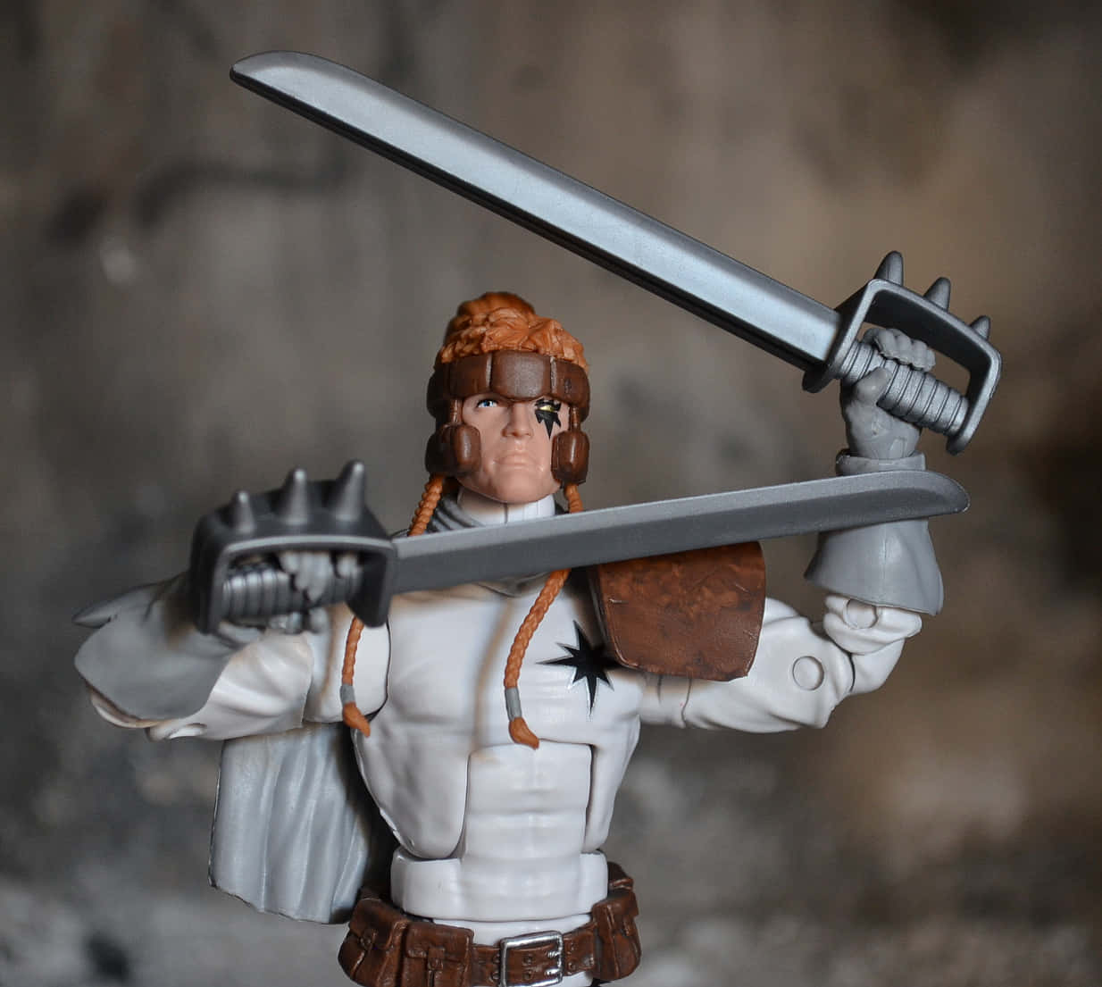 Shatterstar 桌布 壁紙 — 一個細節豐富的動作人偶，描繪一位辮子頭髮、戴眼罩的戰士，在模糊的岩石背景前，以動態戰鬥姿勢手持兩把劍。