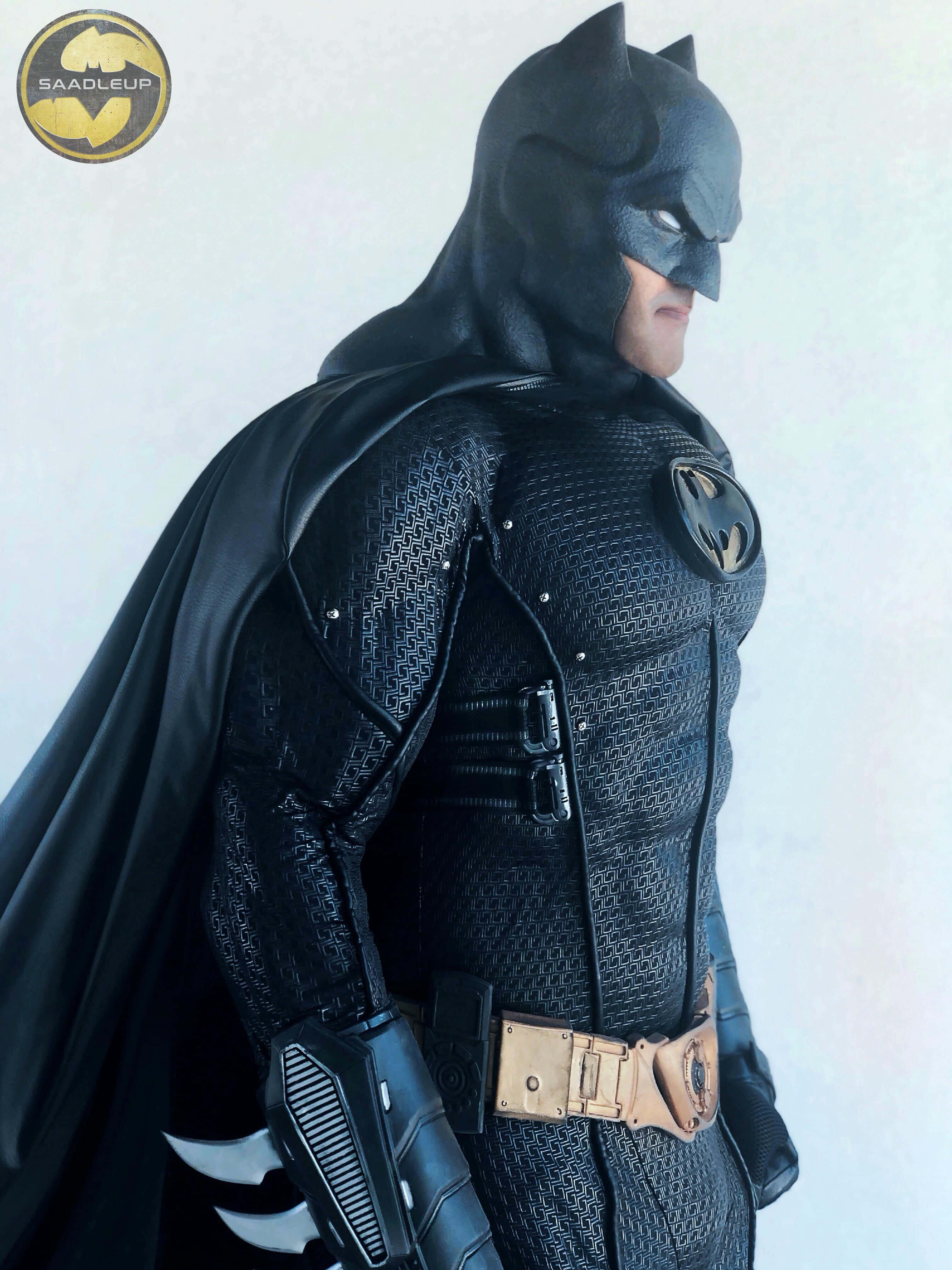 Bat-suit 桌布 壁紙 — 一個細節豐富的蝙蝠俠人偶，身穿帶有紋理的黑色戰衣，配有金色腰帶和披風，在純白色背景前擺出姿勢。