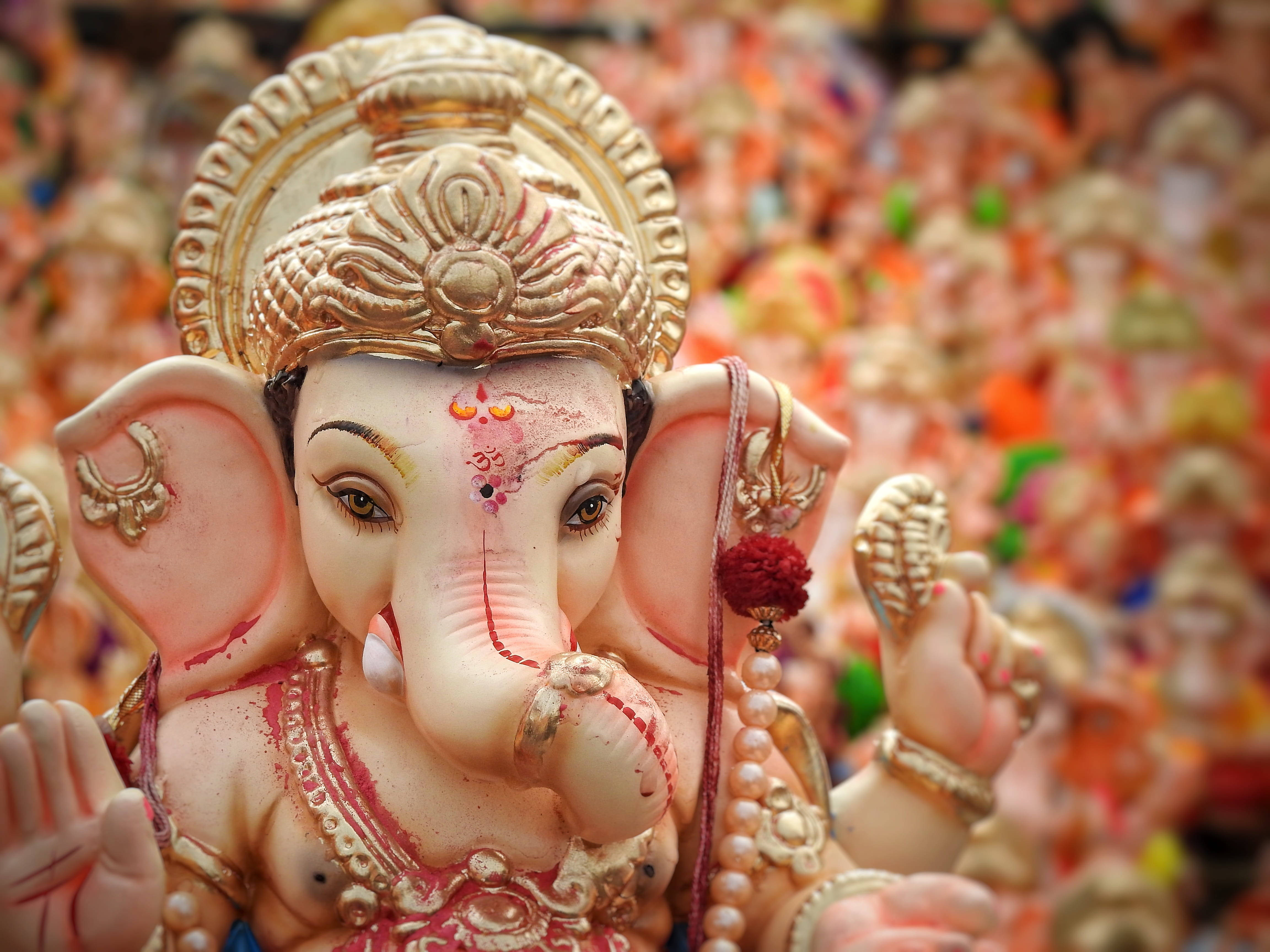 Hindu God 桌布 壁紙 — 一個細節豐富的象神偶像特寫，飾有黃金首飾和紅色標記，周圍是模糊的彩色雕像。