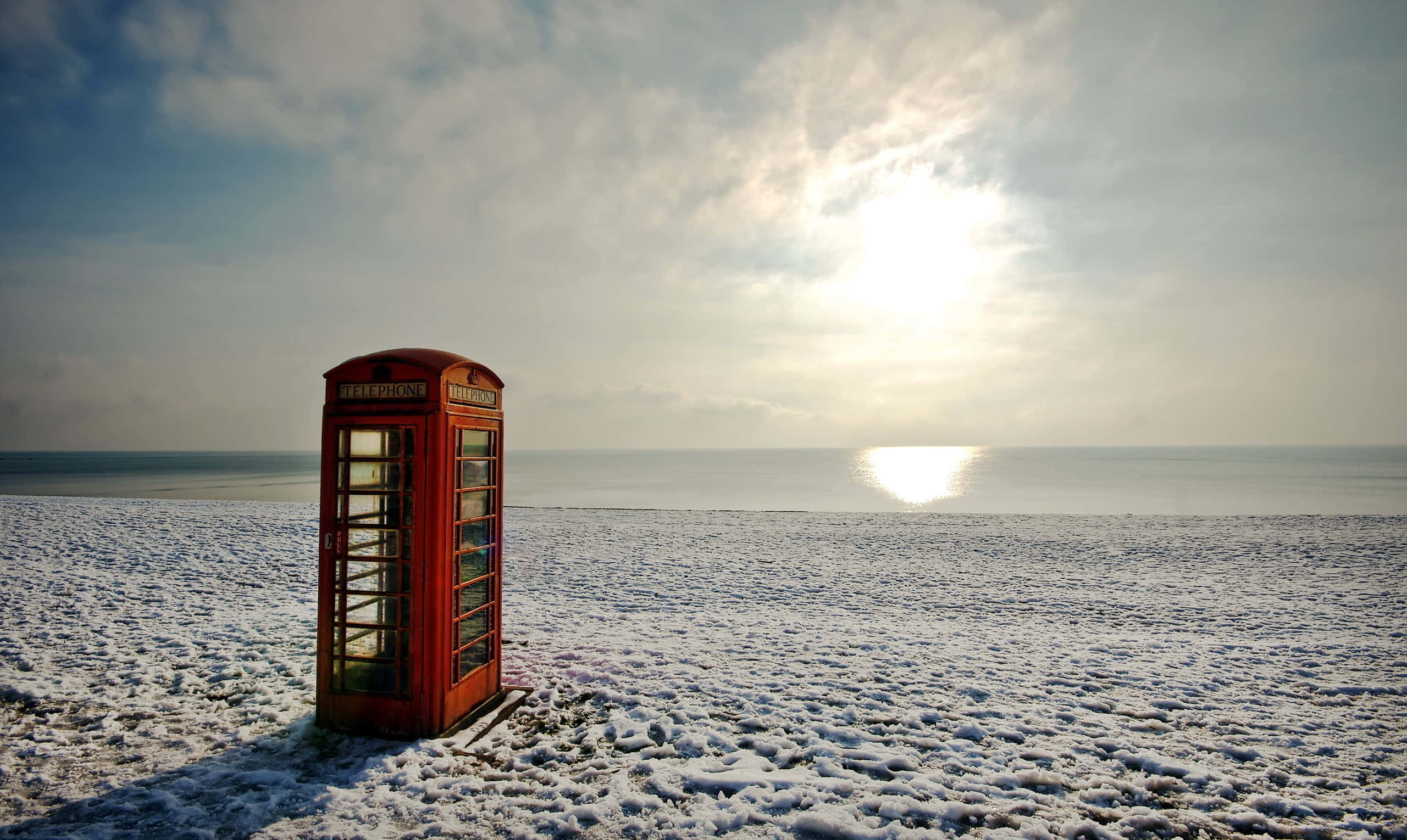 Winter Beach 桌布 壁紙 — 一個經典的紅色英國電話亭獨自矗立在白雪覆蓋的海灘上，太陽在平靜的海面上閃耀。