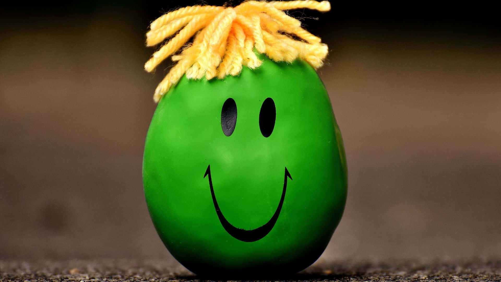 Smiley 桌布 壁紙 — 一個綠色雞蛋，帶有開心的笑臉和黃色毛線頭髮，坐在有紋理的表面上。