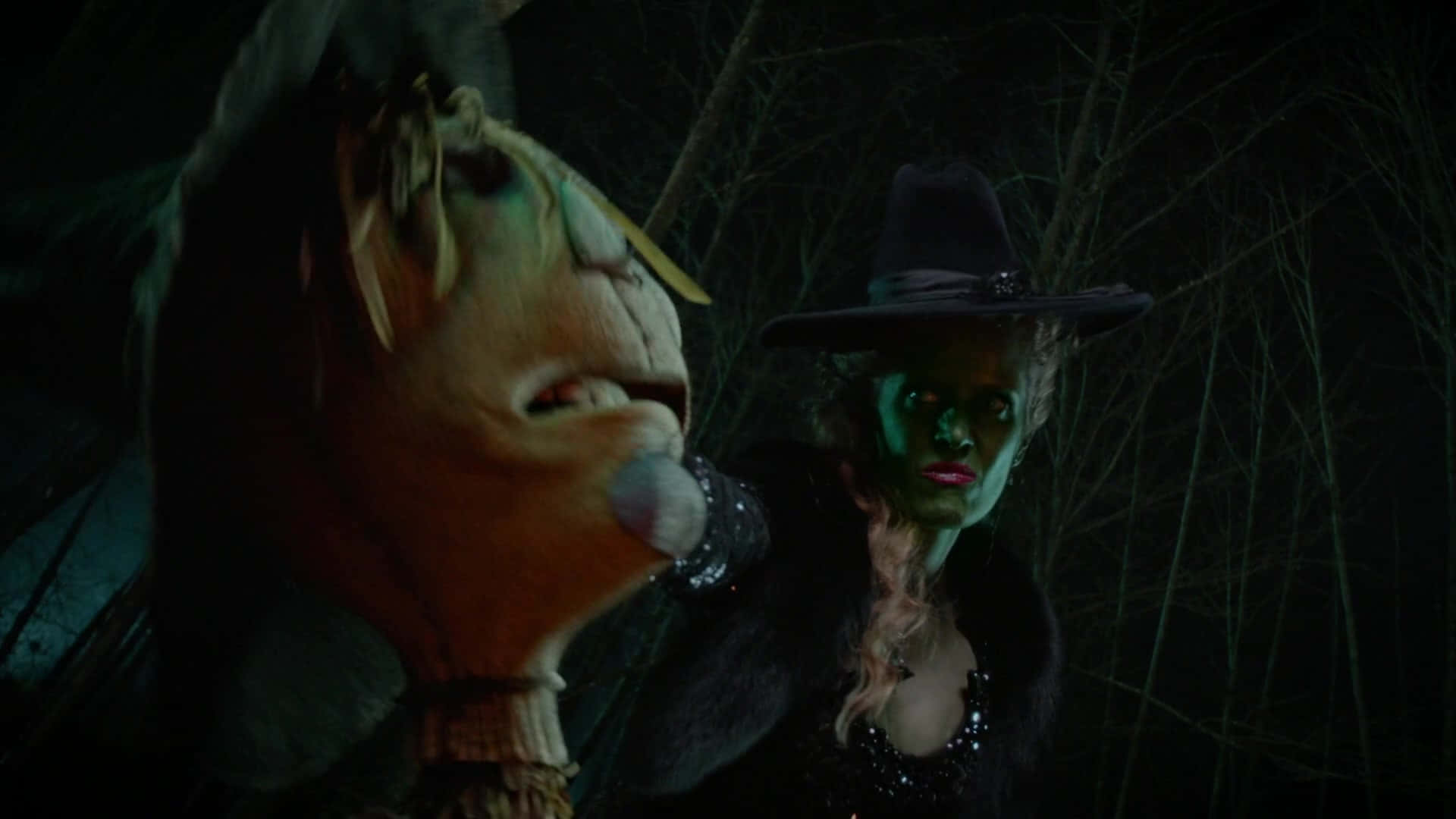 Evil Witch 桌布 壁紙 — 一個綠色面孔、戴黑色帽子的女巫，在夜晚黑暗陰森的森林中站在一個大型木偶頭旁邊。