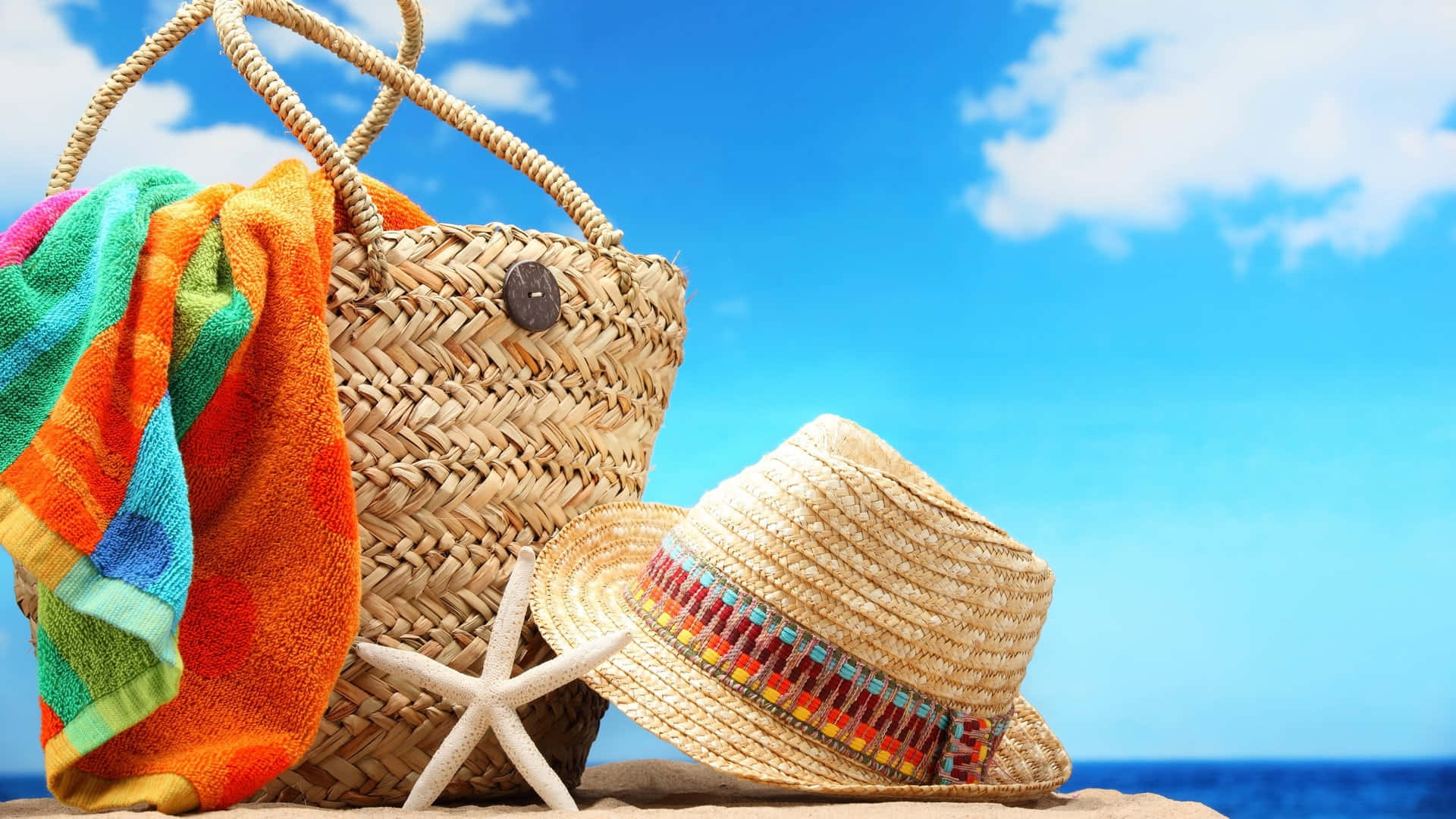 Beach Bag 桌布 壁紙 — 一個編織沙灘袋、一條色彩繽紛的毛巾和一頂草帽放在沙灘上，旁邊有一隻海星，上方是明亮的藍天。