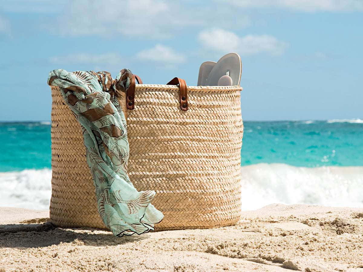 Beach Bag 桌布 壁紙 — 一個編織草沙灘籃，內有圖案圍巾和人字拖，放在金色沙灘上，背後有藍綠色海浪拍打。