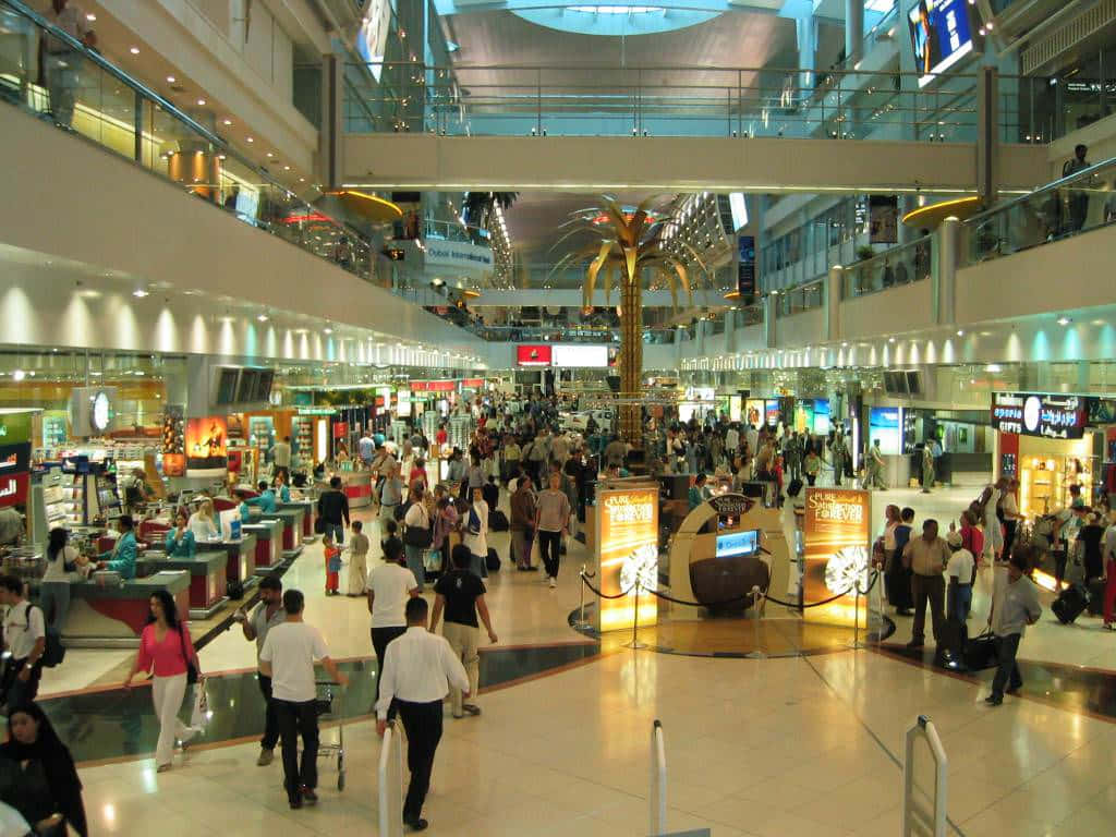 Shopping 桌布 壁紙 — 一個繁忙的多層機場客運大樓，旅客在明亮的燈光下經過商店和資訊亭。