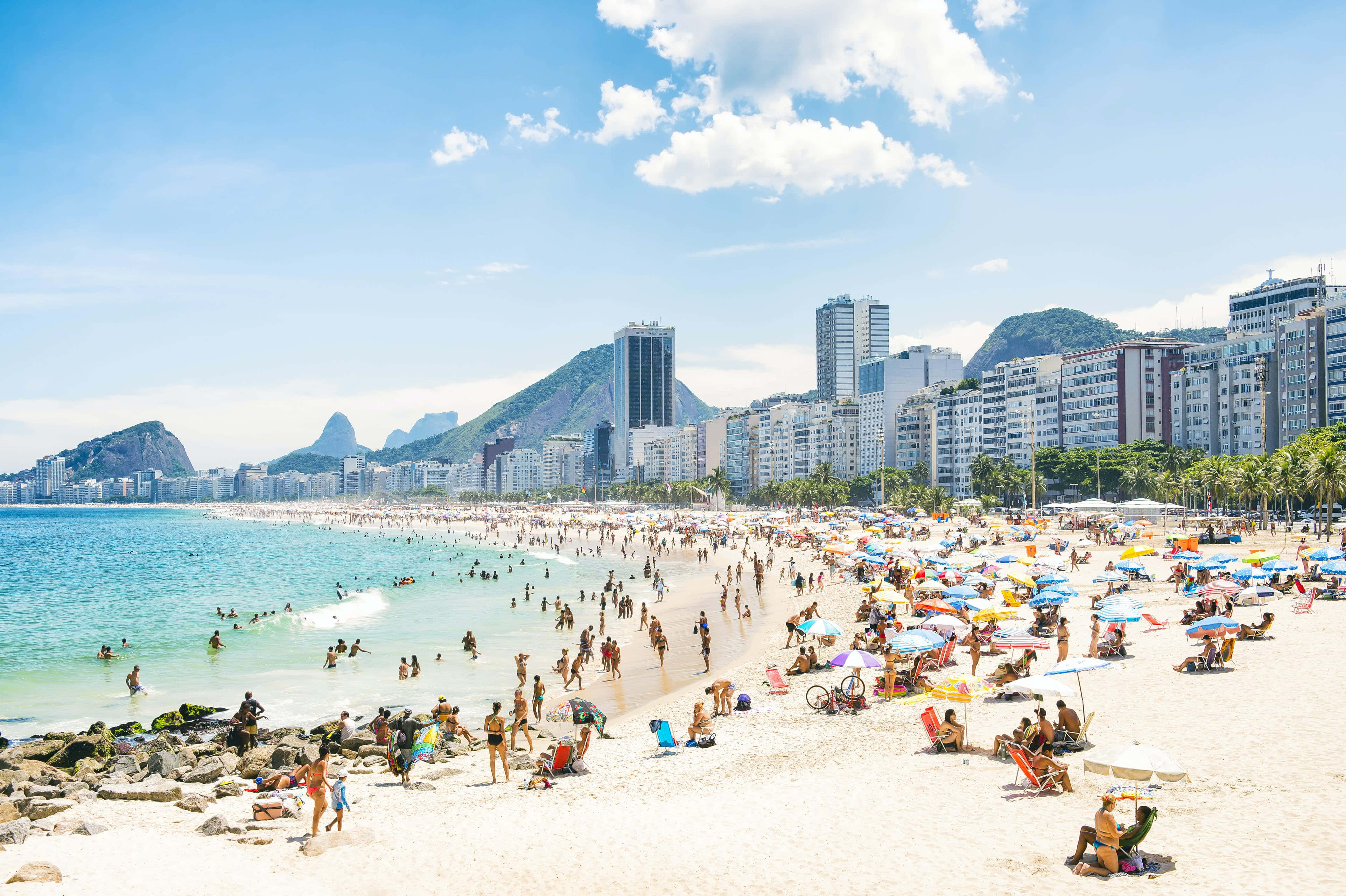 Brazilian Beach 桌布 壁紙 — 一個繁忙的里約海灘，有在彩色遮陽傘下享受日光浴的人們、碧綠的海浪，以及現代建築背後標誌性的山脈。